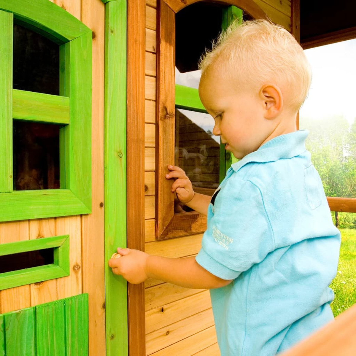 Blondes Kleinkind öffnet eine Holztür eines Spielhauses. Spielhaus ist grün und braun. Blaues Hemd.