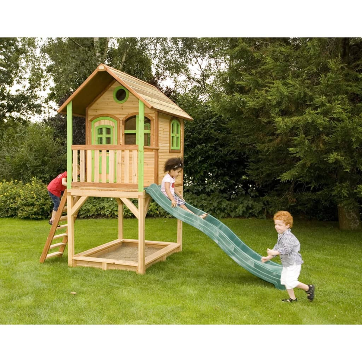 Kinder spielen auf einem Holzspielhaus mit Rutsche und Leiter in einem Rasen mit Bäumen.