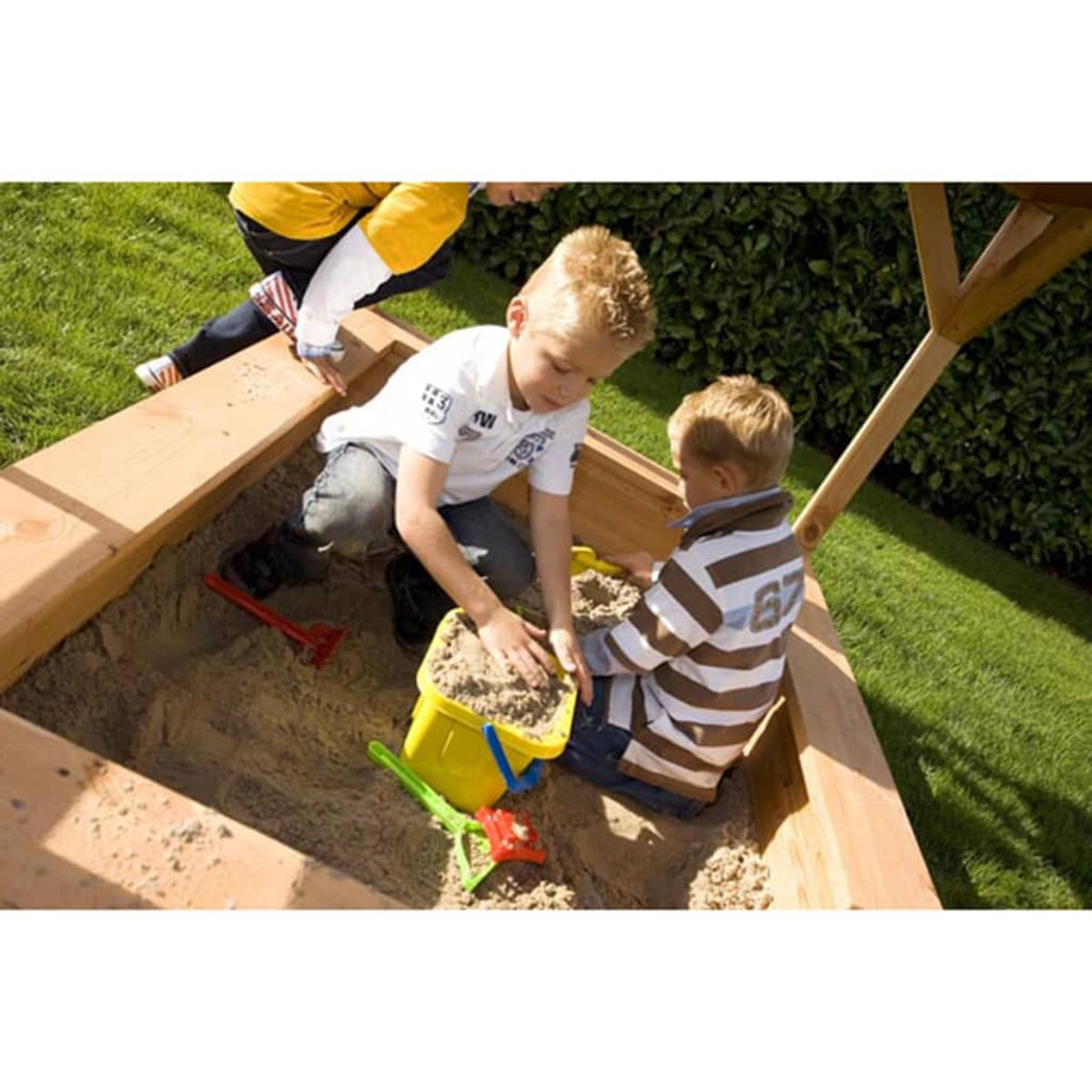 Kinder spielen in einem Sandkasten mit einem gelben Eimer, Schaufeln und Rasen im Hintergrund.