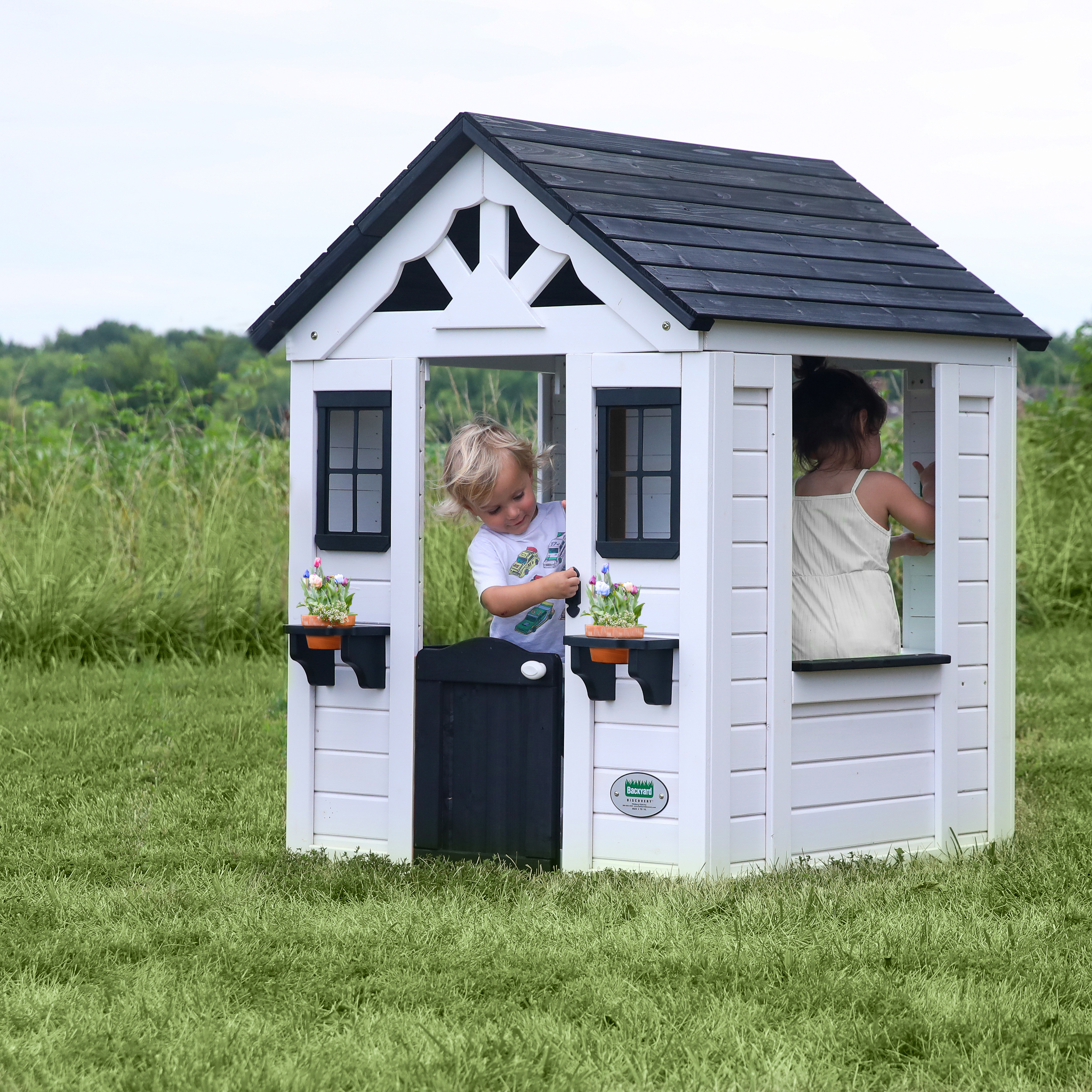 Weißes Spielhaus mit schwarzem Zierrat. Zwei Kinder schauen aus Fenstern und Tür, auf grünem Gras.