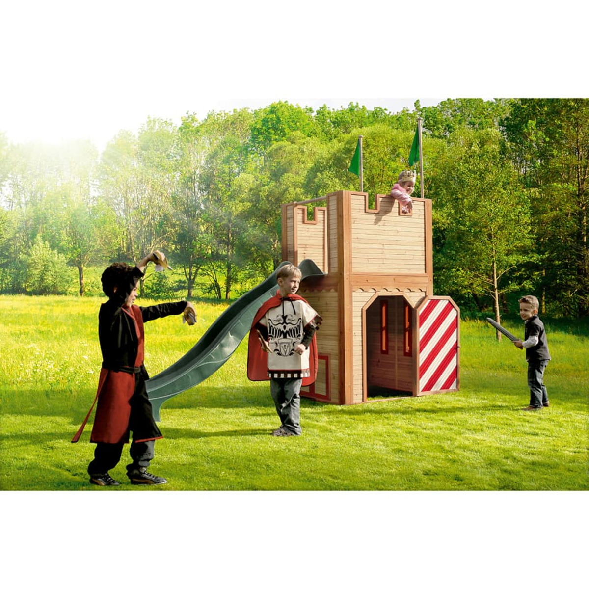 Kinder spielen in Holzburg mit Rutsche. Grünes Gras, Bäume im Hintergrund.