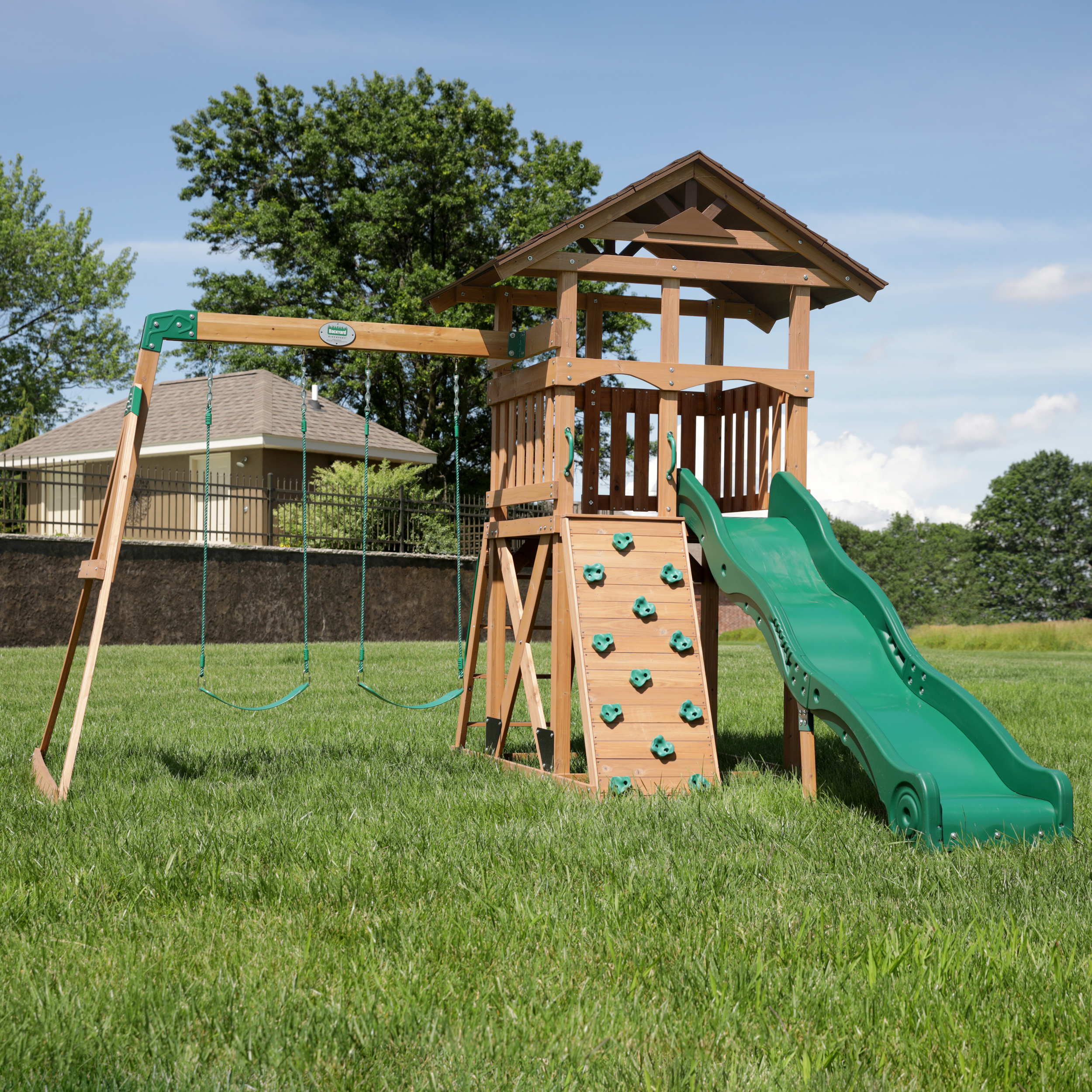 Ein Spielplatz aus Holz mit Schaukeln, Kletterwand, Rutsche und Turm, auf Gras.