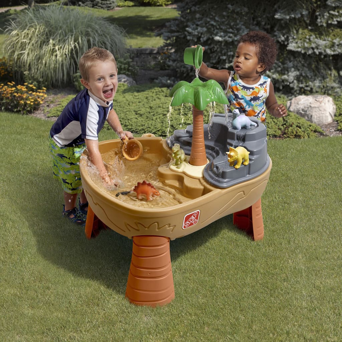 Zwei Kinder spielen mit Wasser in einem wassertisch mit Dinosaurier-Motiv auf grünem Gras.