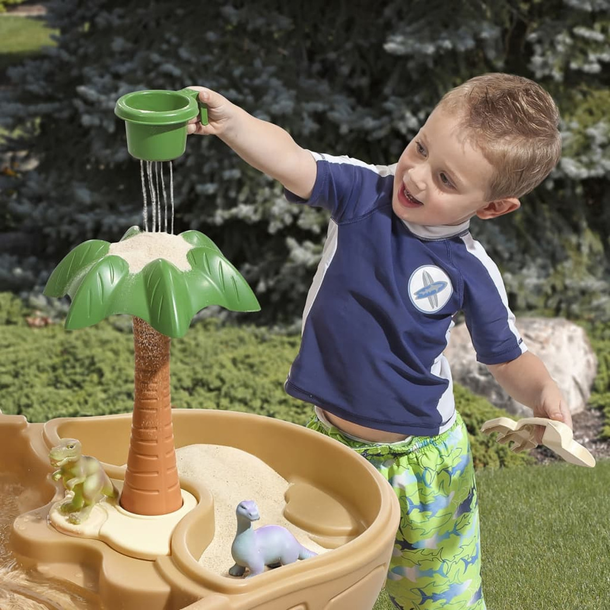 Ein kleiner Junge spielt mit einem Sand- und Wassertisch und gießt Wasser aus einem grünen Becher.
