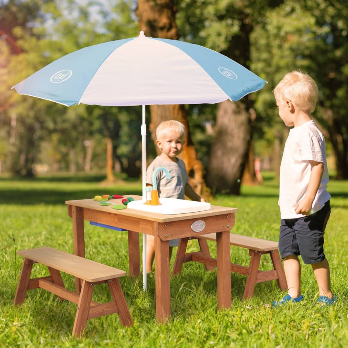 Zwei Kinder an einem Holztisch mit Sonnenschirm. Grünes Gras und Bäume im Hintergrund.