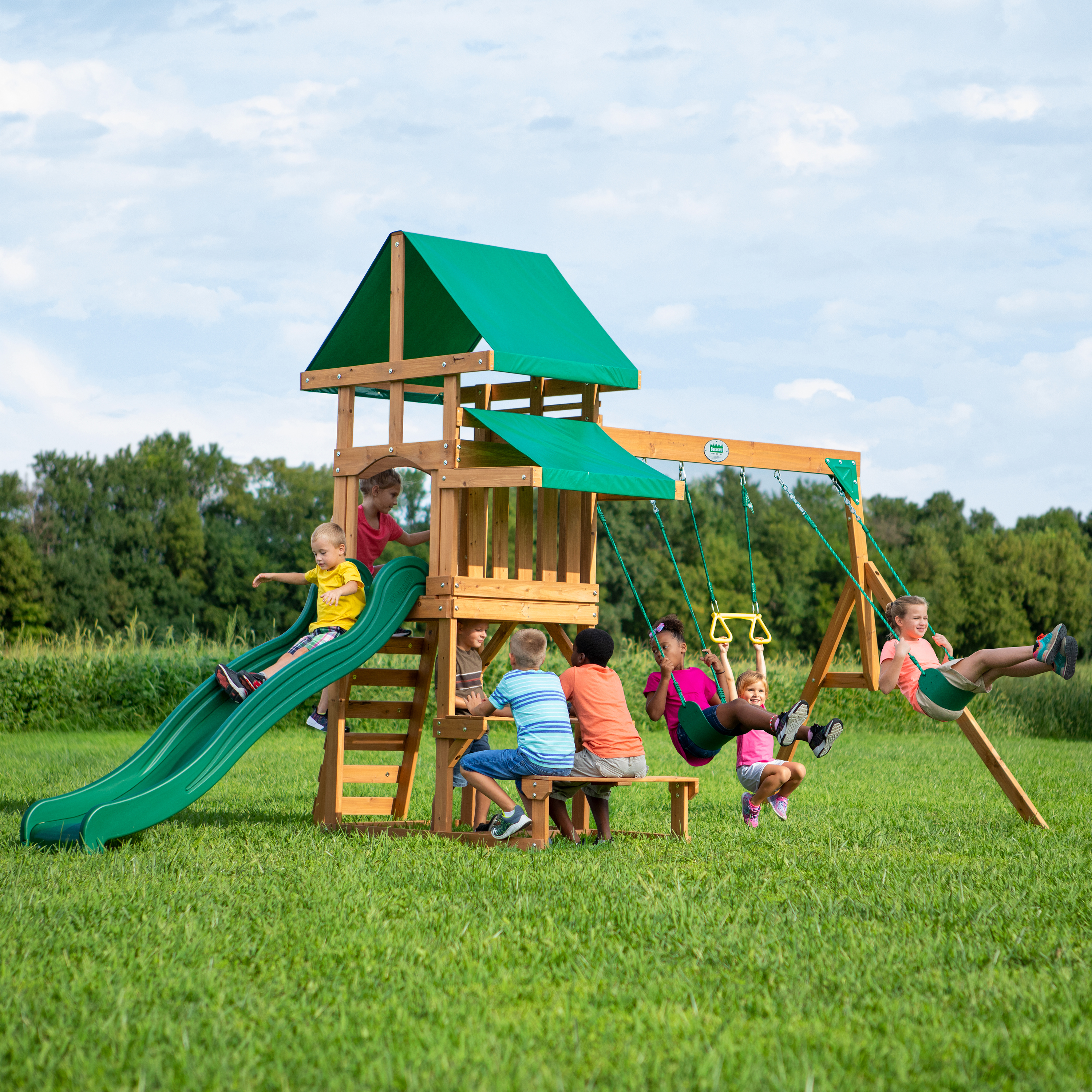 Kinder spielen auf einem Holzspielplatz. Grüne Rutsche, Schaukeln und ein kleines Spielhaus. Grüner Rasen.