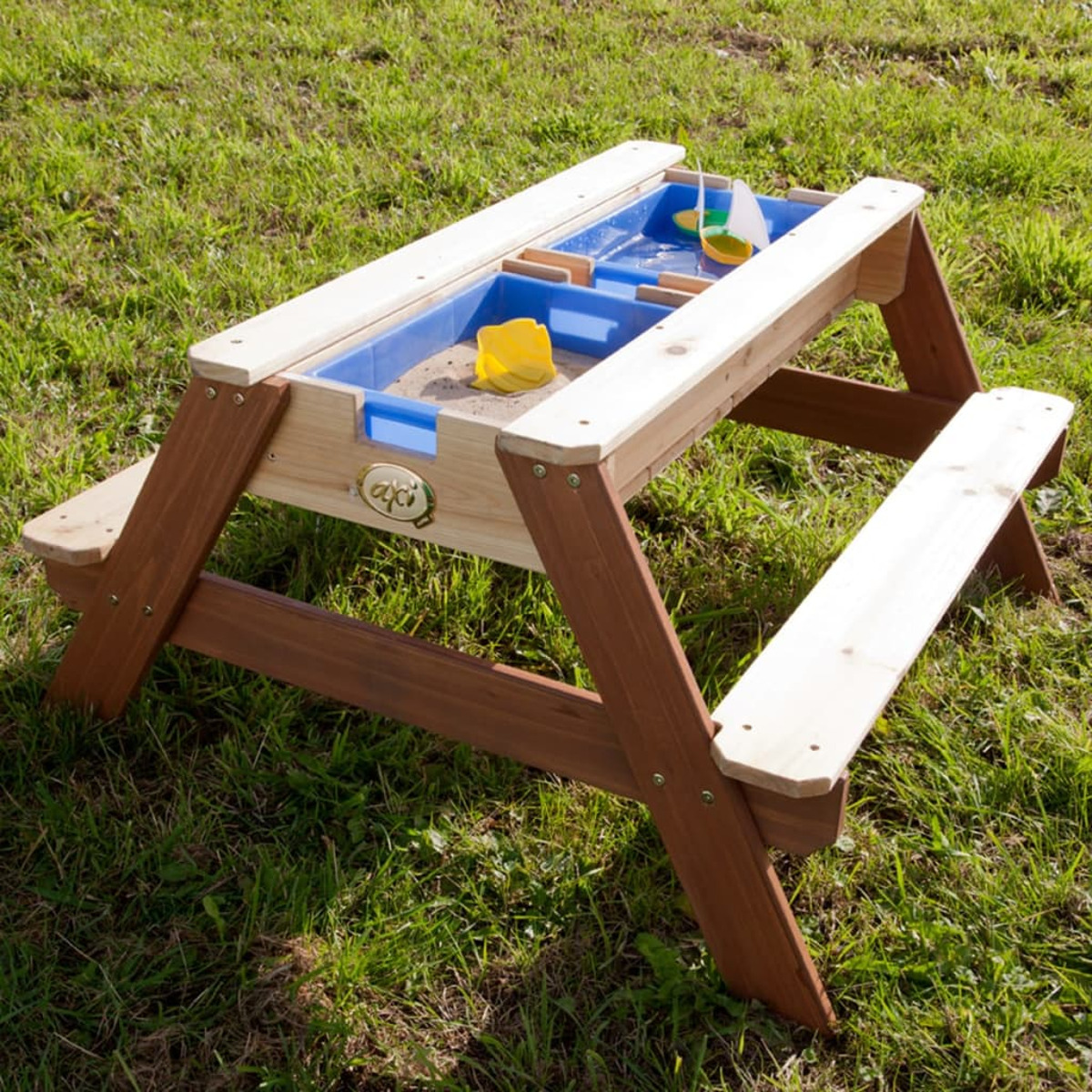Ein hölzerner Picknicktisch mit blauem Becken und gelbem Spielzeug auf grünem Gras. Der Tisch hat zwei Bänke.