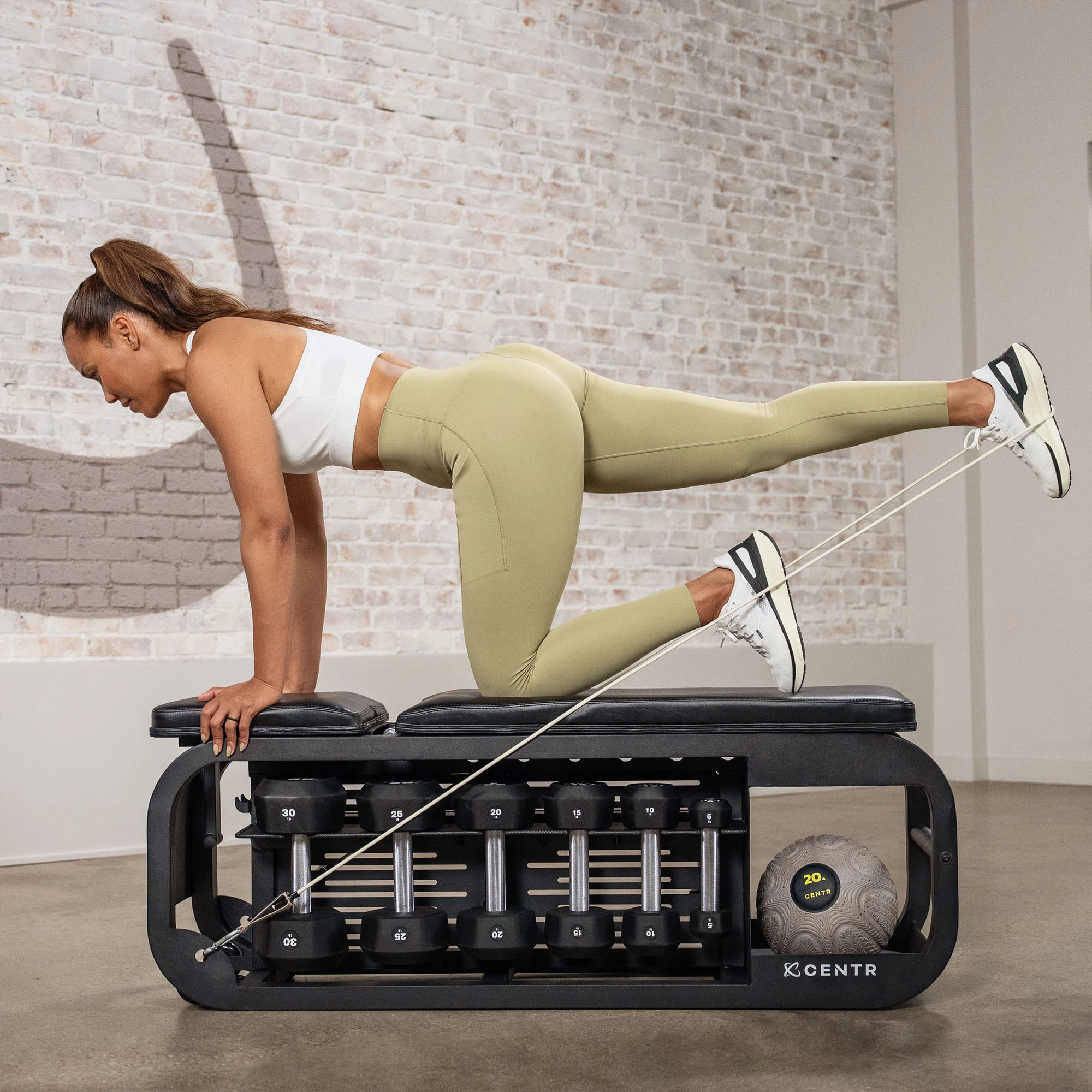 Woman doing exercise on a bench. She is using a resistance band, with weights and ball in the background.