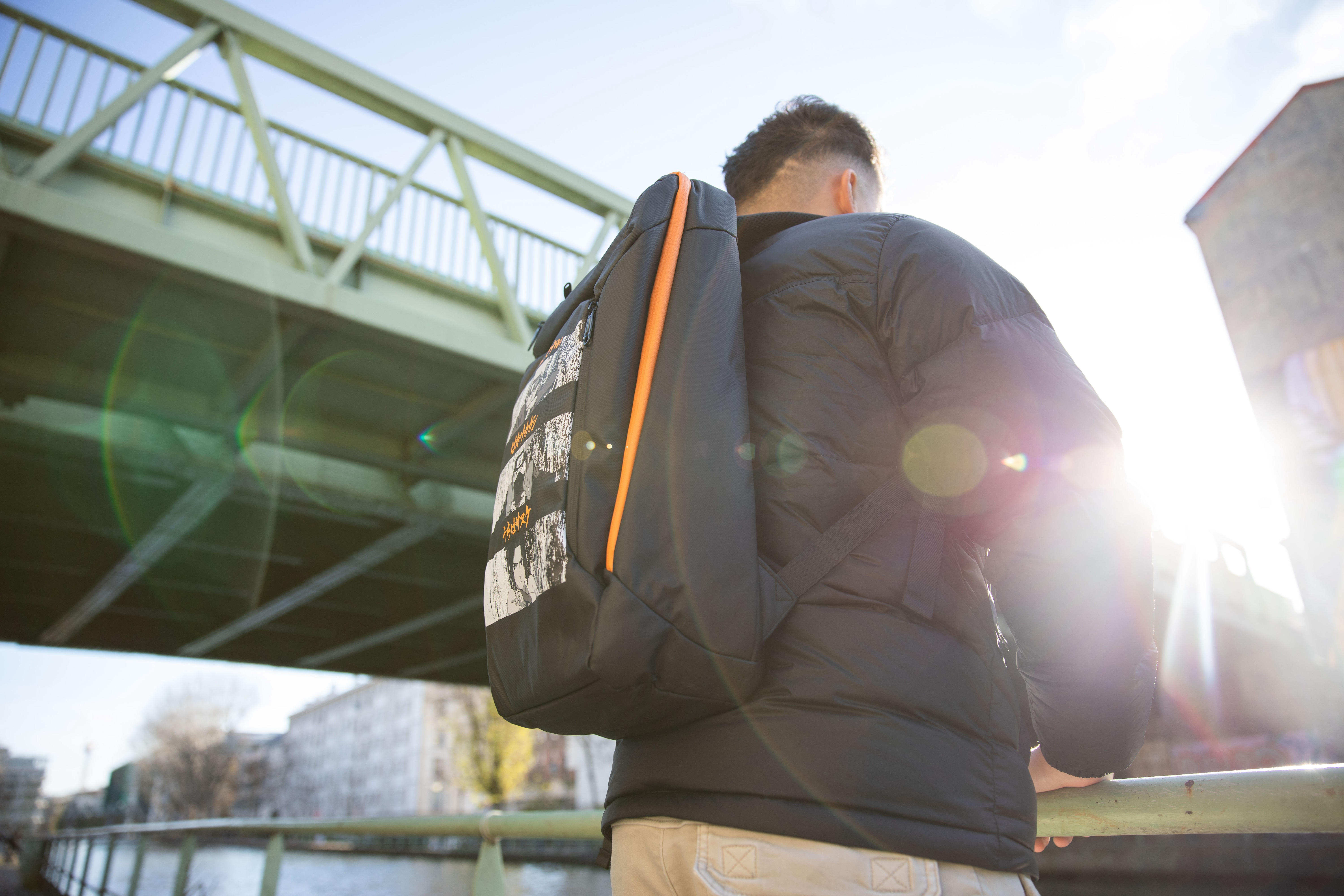 Eine Person mit Rucksack steht in der Nähe einer Brücke. Die Sonne scheint. Der Rucksack ist schwarz und orange.