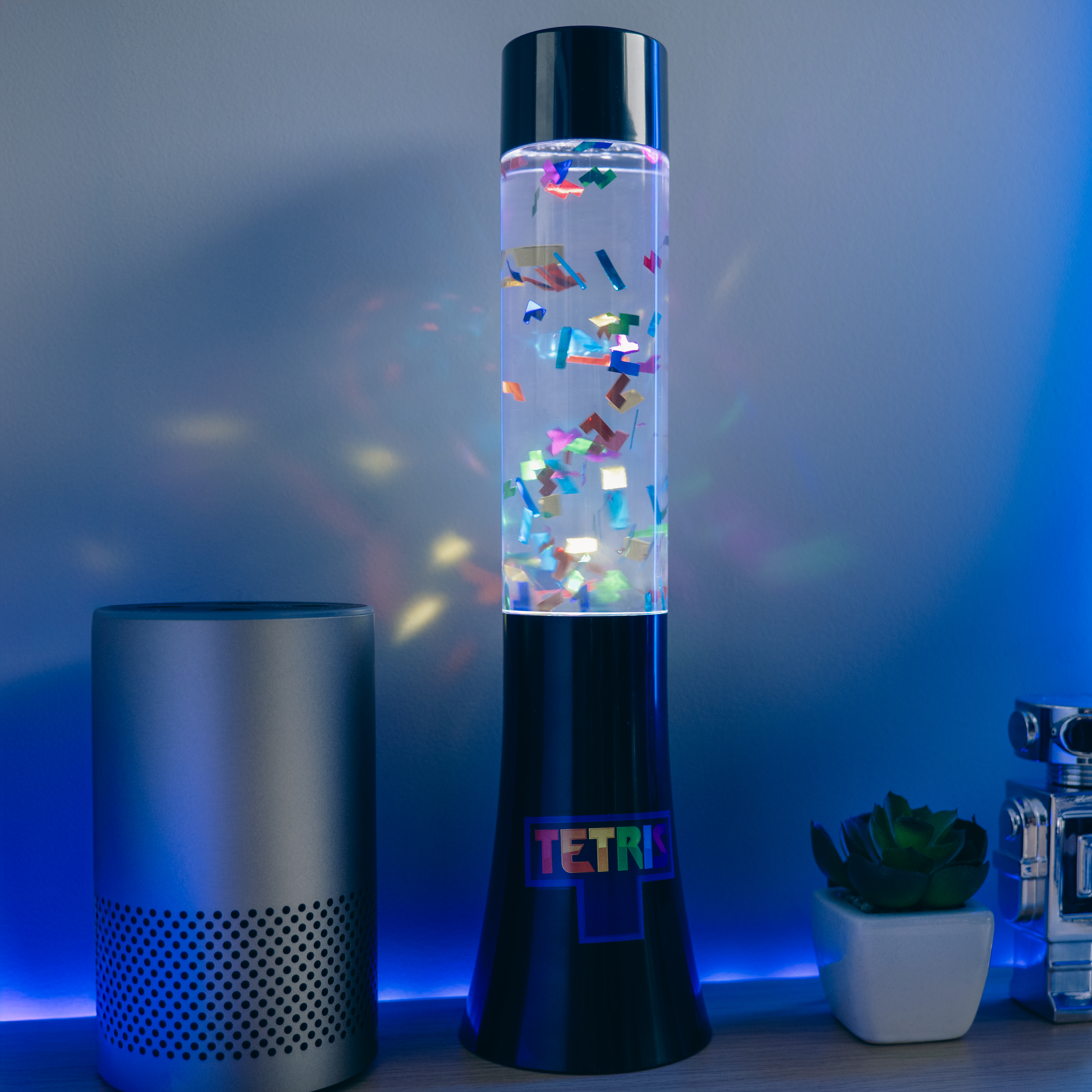 Tetris-lamp, kleurrijke blokken in een heldere cilinder. Zilveren speaker en een kleine plant.