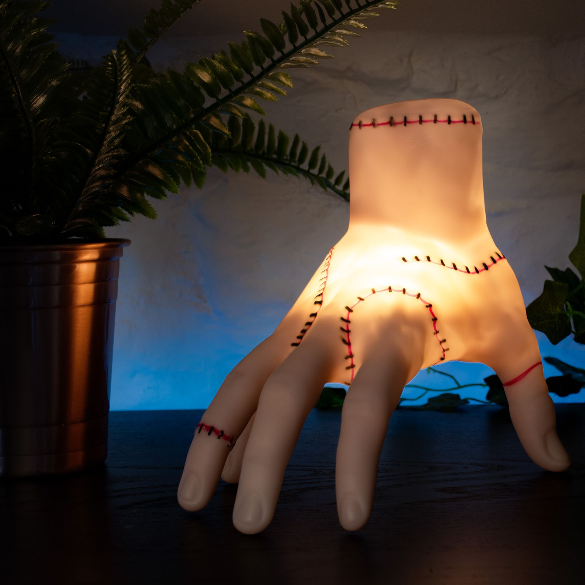 Une lampe en forme de main, avec des coutures rouges. Une fougère en pot à gauche. Fond bleu flou et mur blanc.