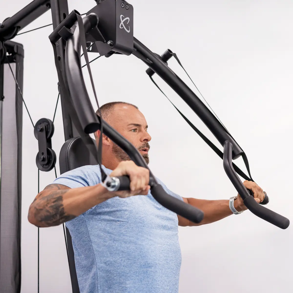 Ein Mann benutzt eine Brustpresse im Fitnessstudio. Er hat ein Tattoo und schaut nach vorne.
