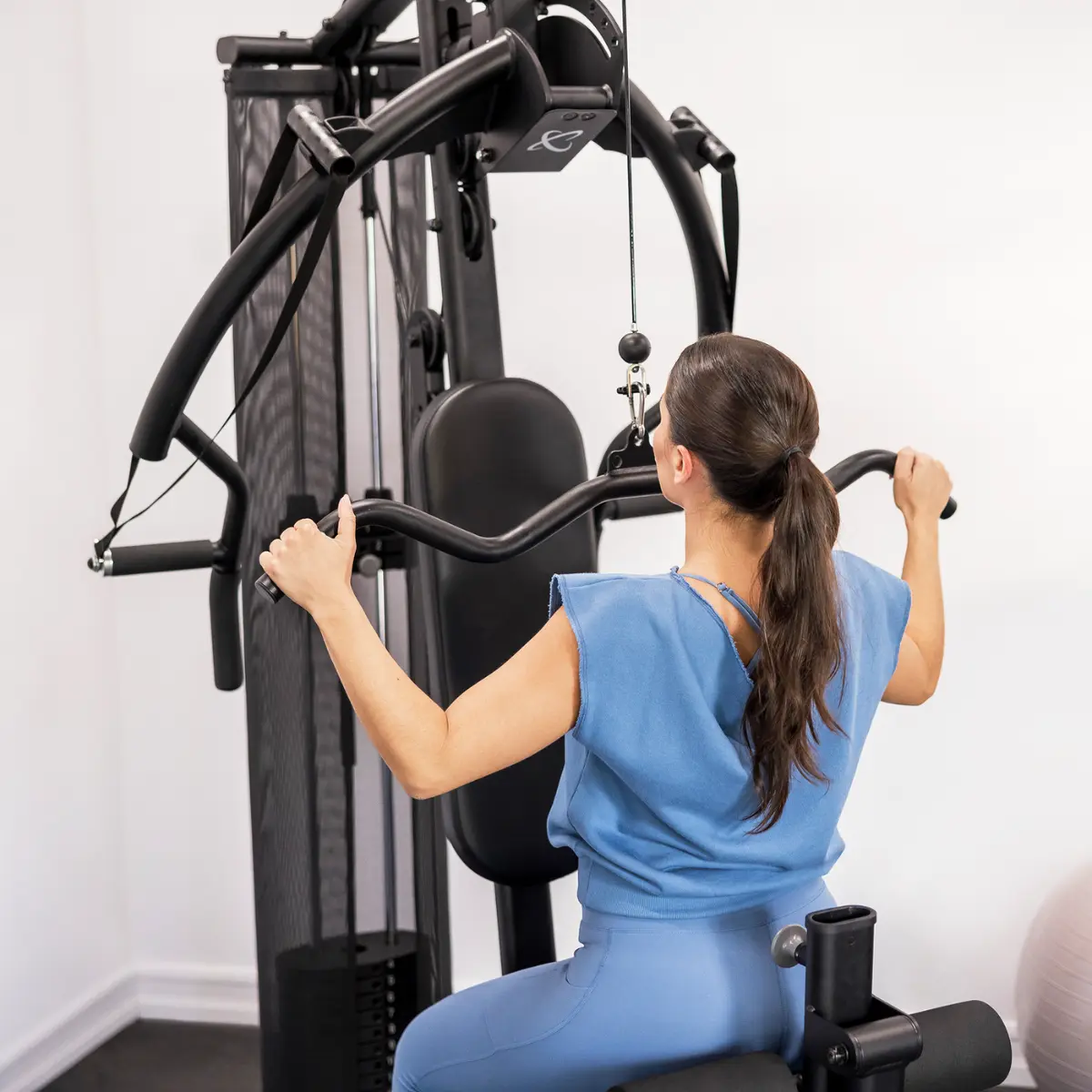 Eine Frau benutzt ein Fitnessgerät in einem Fitnessstudio. Sie trägt blaue Kleidung.