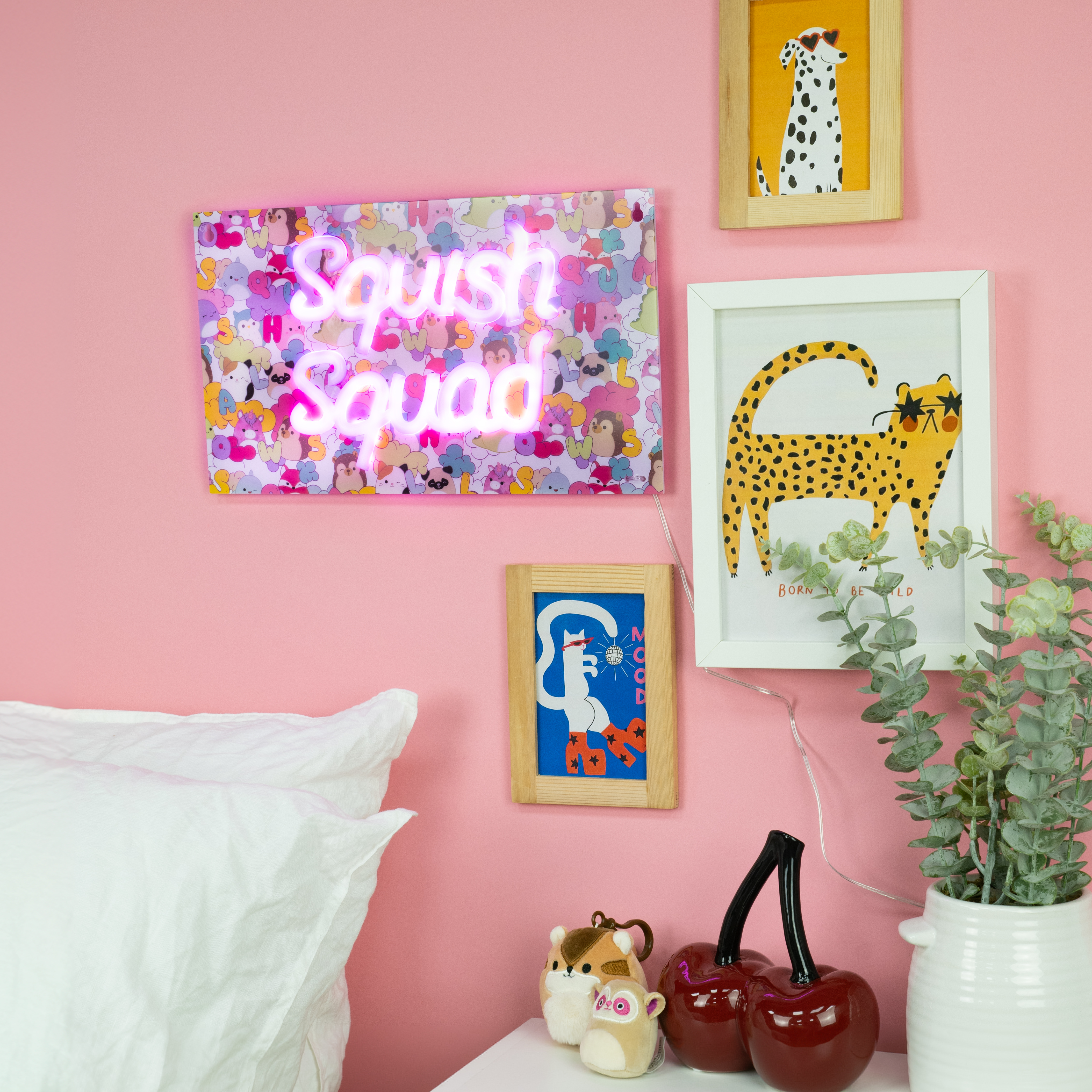 Roze muur met ingelijste kunst en een neonbord met 'Squish Squad'. Een bed en decor staan ervoor.