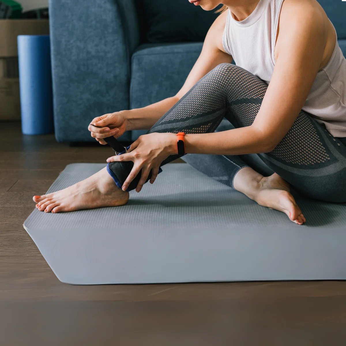Vrouw doet een enkelgewicht aan. Grijze legging en een licht tanktopje.