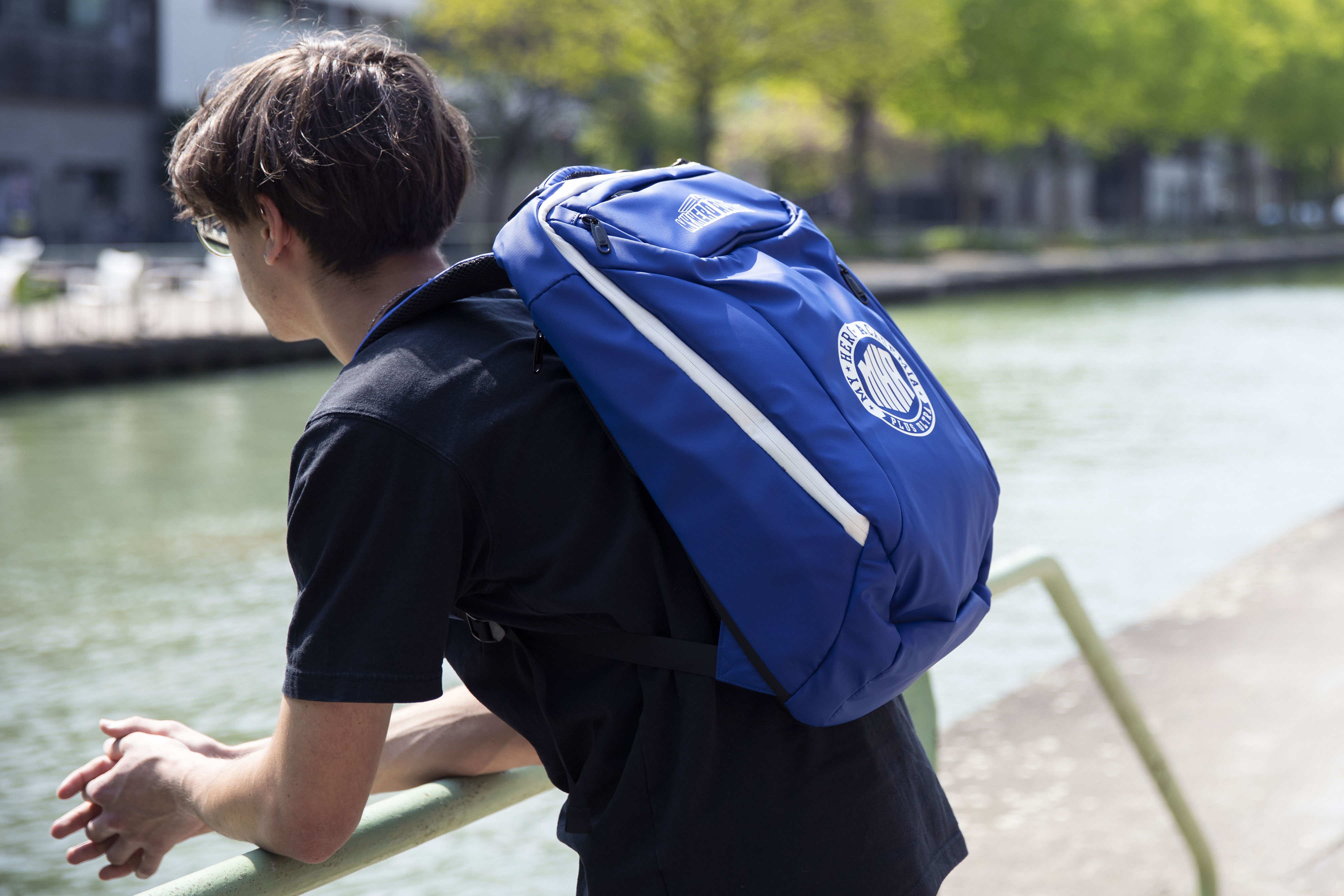 Eine Person mit blauem Rucksack geht an einem Kanal entlang. Die Tasche hat ein Logo.