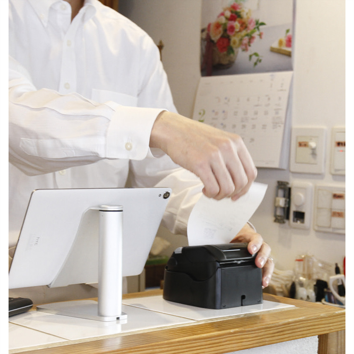 A person taking a receipt from a black printer. A tablet is to the left. White background.