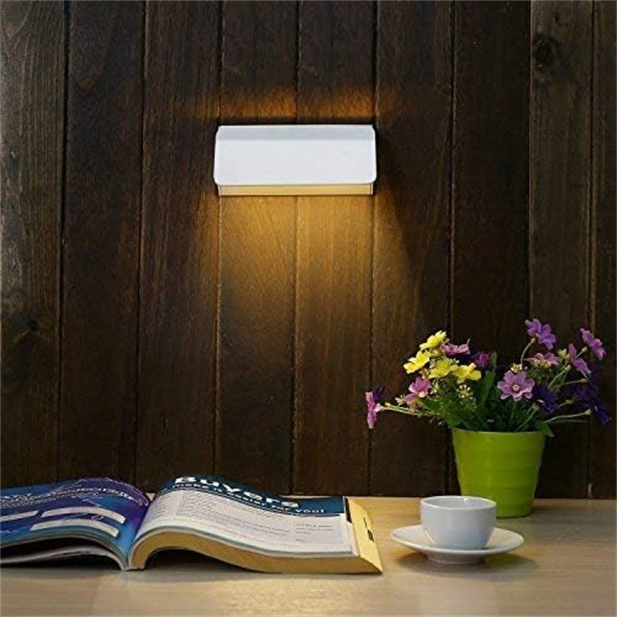 Eine Wandleuchte beleuchtet eine Holzwand, mit Buch, Blumen und einer Tasse.