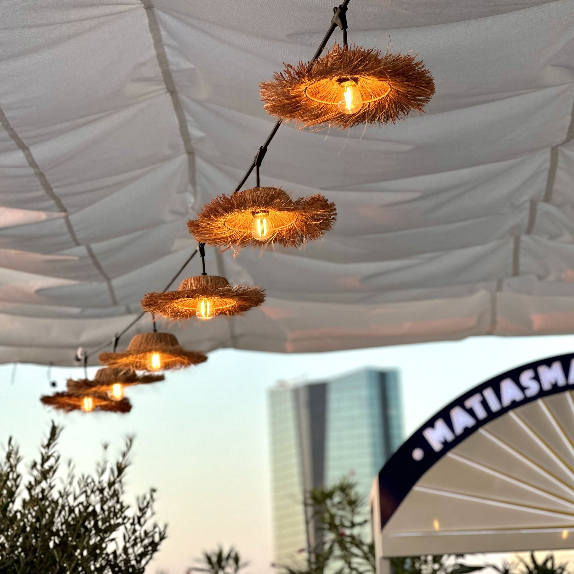 Lichter, die von einem Baldachin hängen. Die Lichter sind kreisförmig, mit strohartigen Schirmen. Ein Gebäude im Hintergrund.