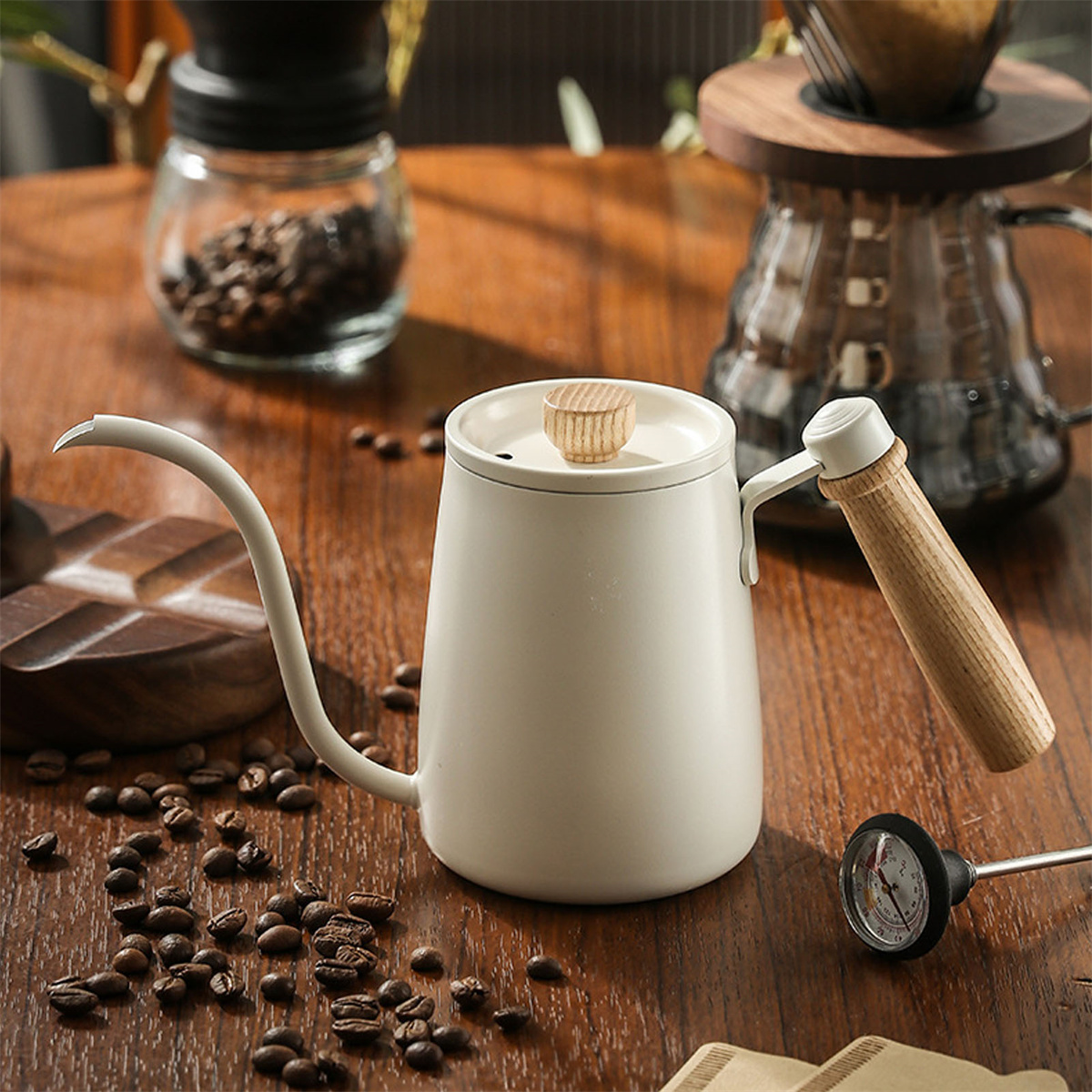 Weißer Schwanenhalskessel auf einem Holztisch mit Kaffeebohnen. Ein Thermometer daneben. Heller Hintergrund.