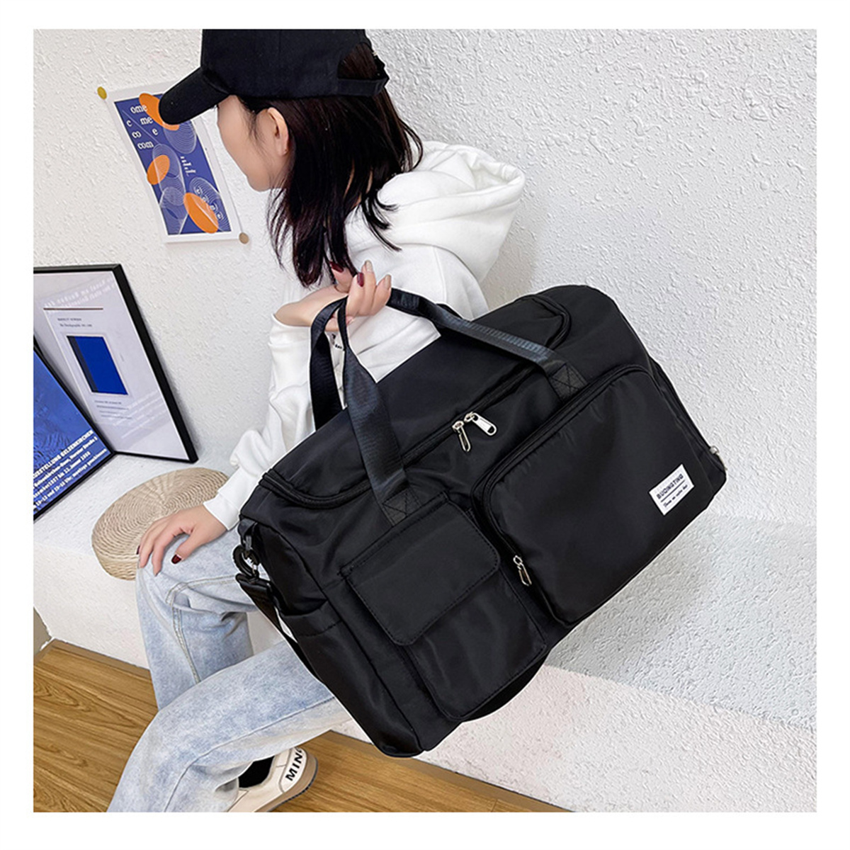 Schwarze Reisetasche mit Taschen auf dem Schoß einer Frau. Sie trägt einen weißen Kapuzenpullover und Jeans.