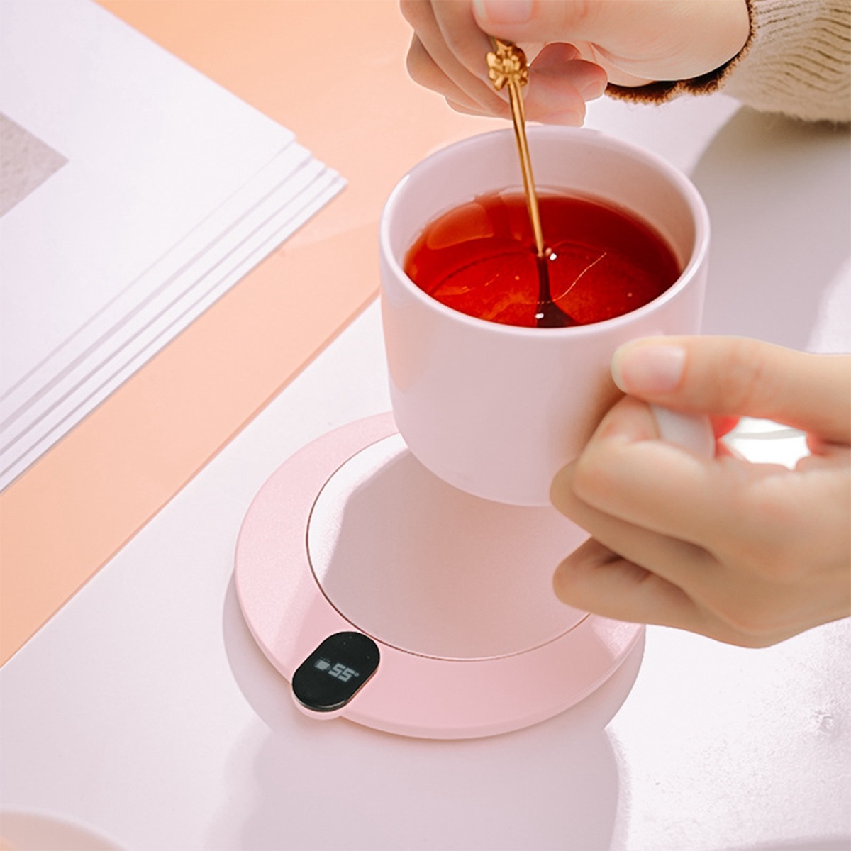 Eine Person hält einen rosafarbenen Becher auf einem beheizten rosa Untersetzer. Ein Löffel im Becher. Weißer Tisch.