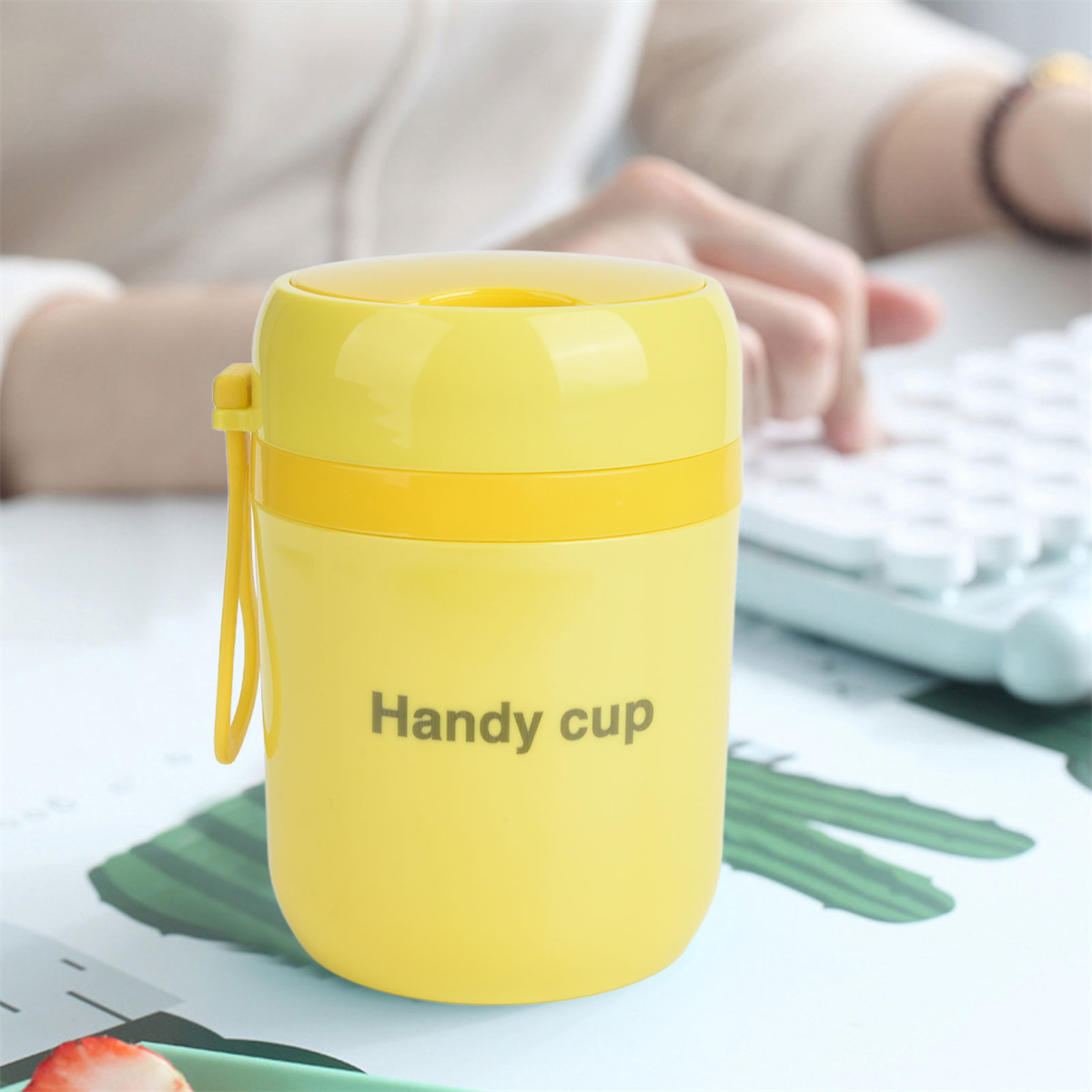 Gelber 'Handy Cup' mit Griff. Eine Hand und Tastatur sind unscharf im Hintergrund.
