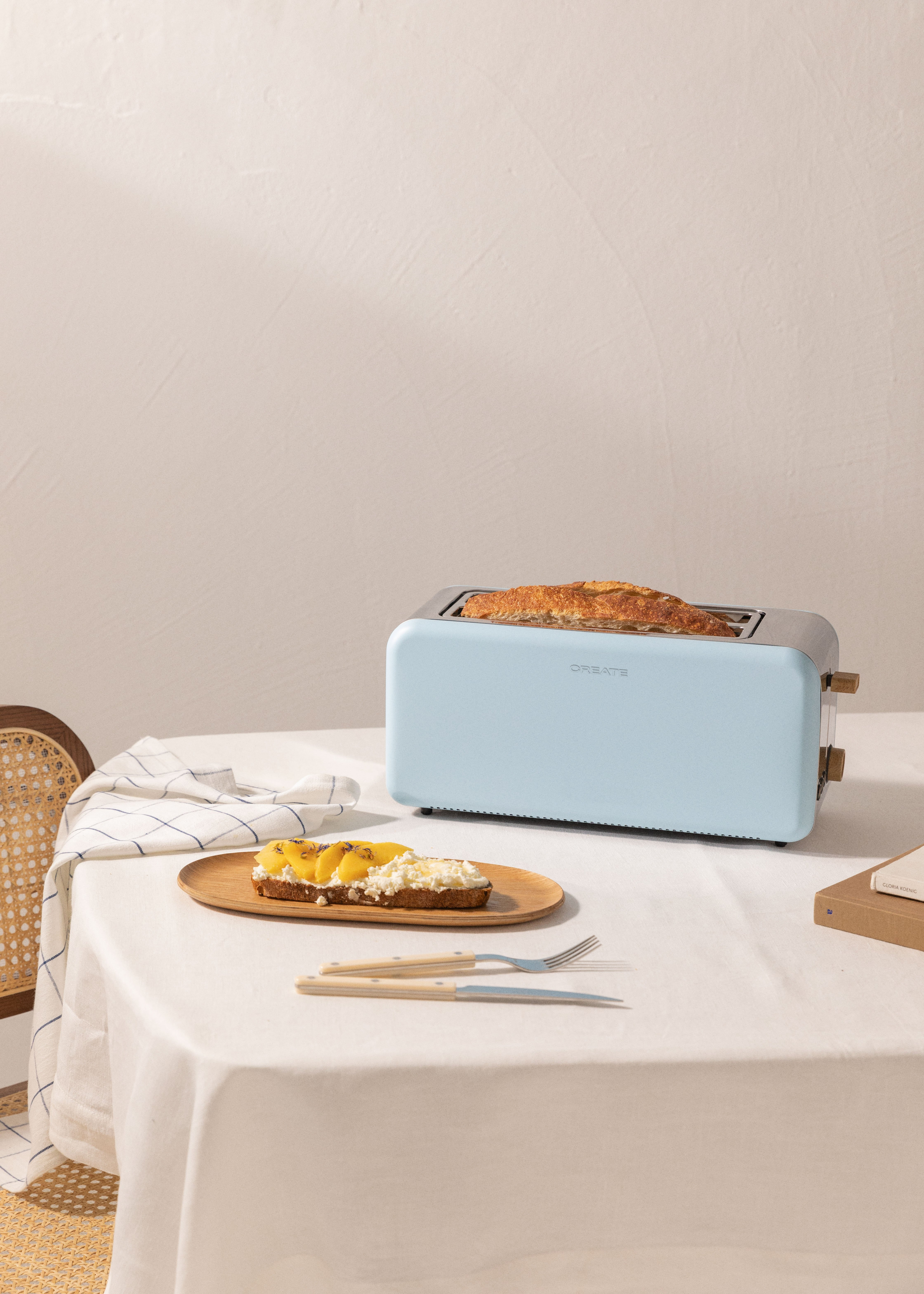 Una tostadora azul claro sobre una mesa blanca, con tostadas, una bandeja de madera y un mantel blanco.
