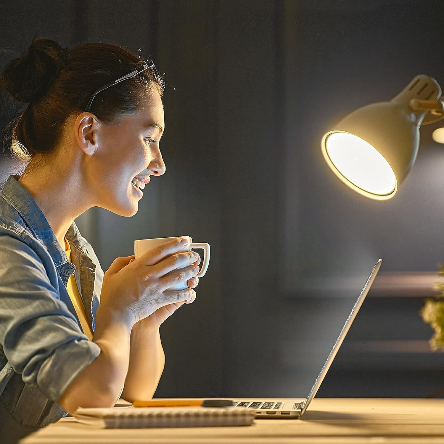 Eine Frau in einem Jeanshemd lächelt, während sie auf einen Laptop blickt und eine Tasse Kaffee unter einer Lampe hält.