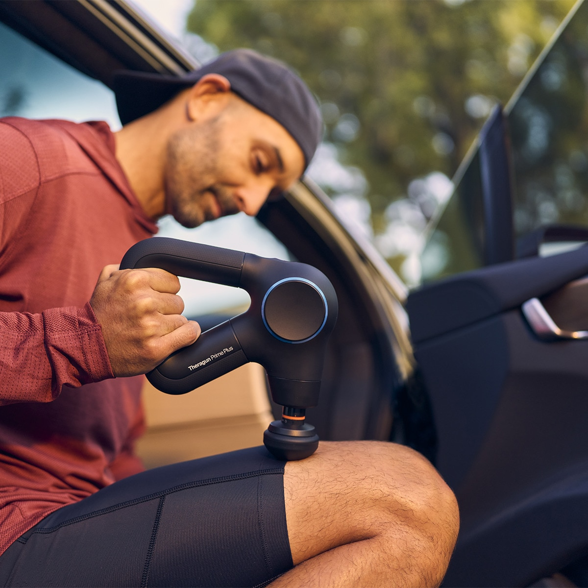 Mężczyzna używa czarnego pistoletu do masażu na udzie w samochodzie. Czarne i niebieskie akcenty.