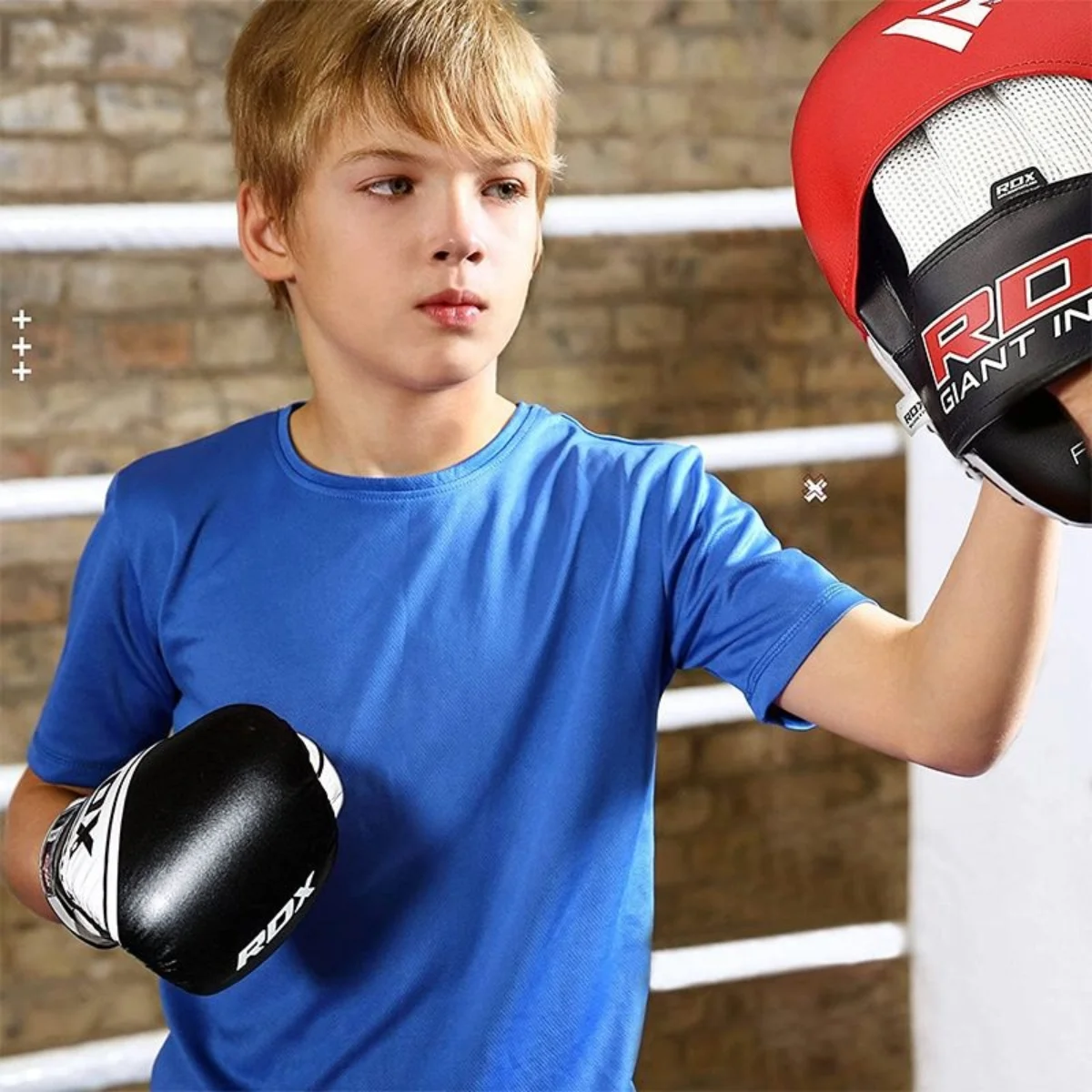 A boy wearing a blue shirt and boxing gloves is holding a training pad.