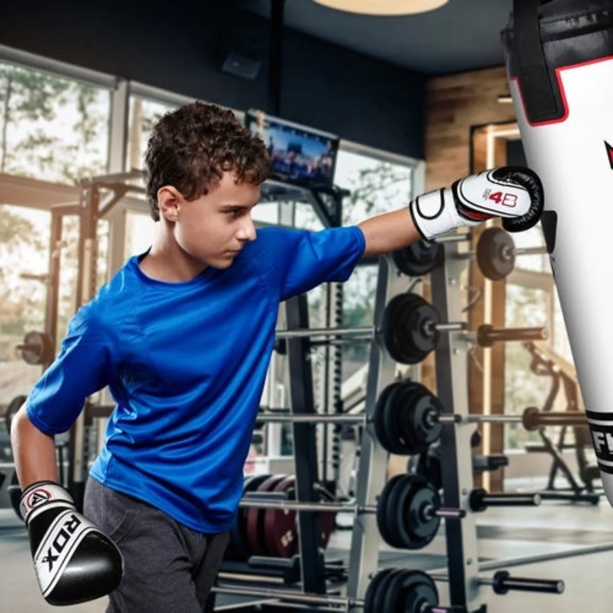 Young boy boxing, wearing blue shirt and gloves, hitting a punching bag in a gym setting.