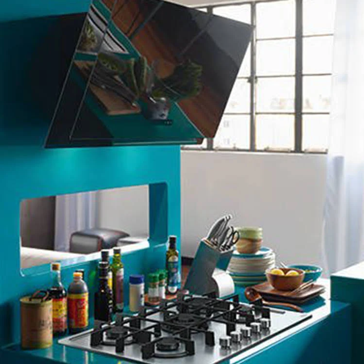 Cucina moderna con pareti turchesi. Piano cottura nero, piano di lavoro in acciaio inox, bottiglie e specchio sospeso.