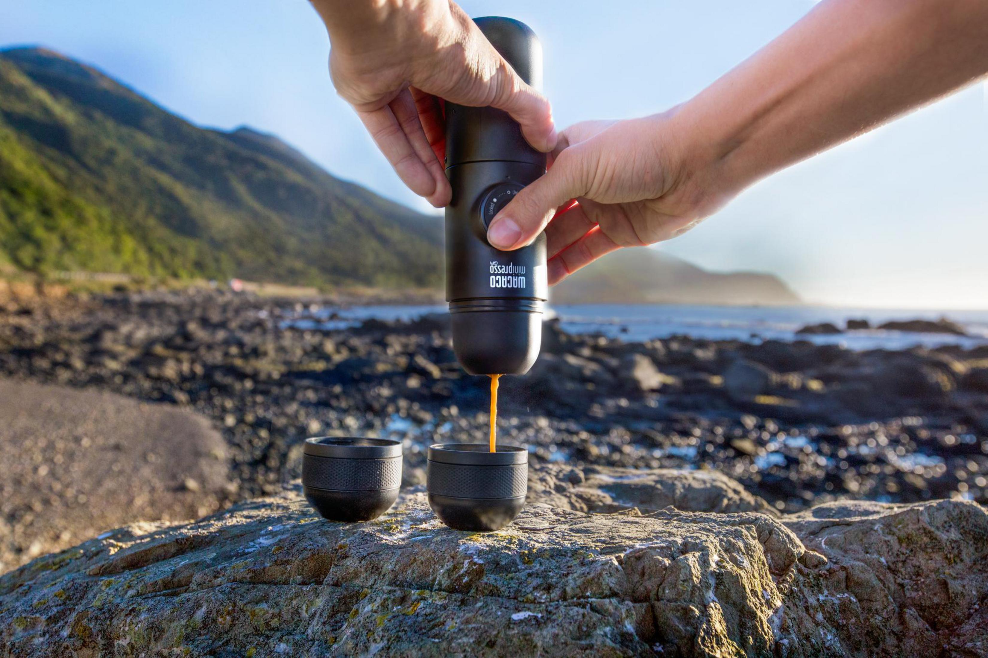 Hände gießen Espresso aus einem tragbaren Kaffeezubereiter in zwei Tassen an einem felsigen Strand am Meer.