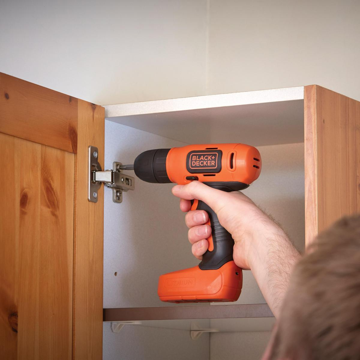 Osoba używająca wiertarki do montażu zawiasu na drewnianej szafce, wewnątrz na białej ścianie.
