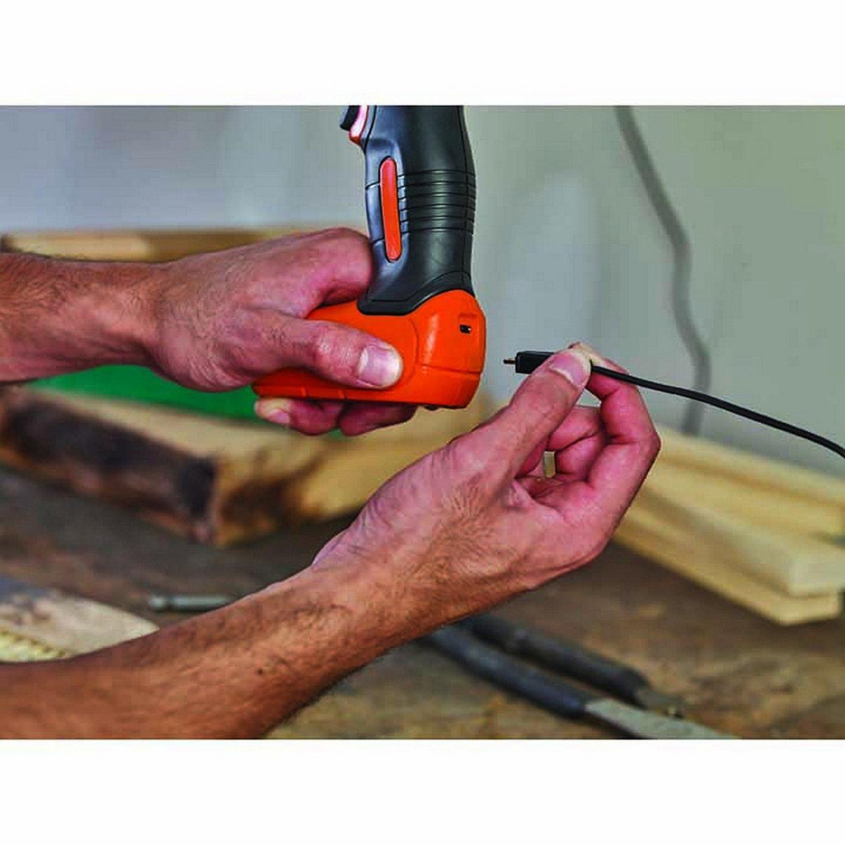 Dłonie trzymające czarno-pomarańczowe narzędzie, podłączające czarny kabel. Drewniana powierzchnia.