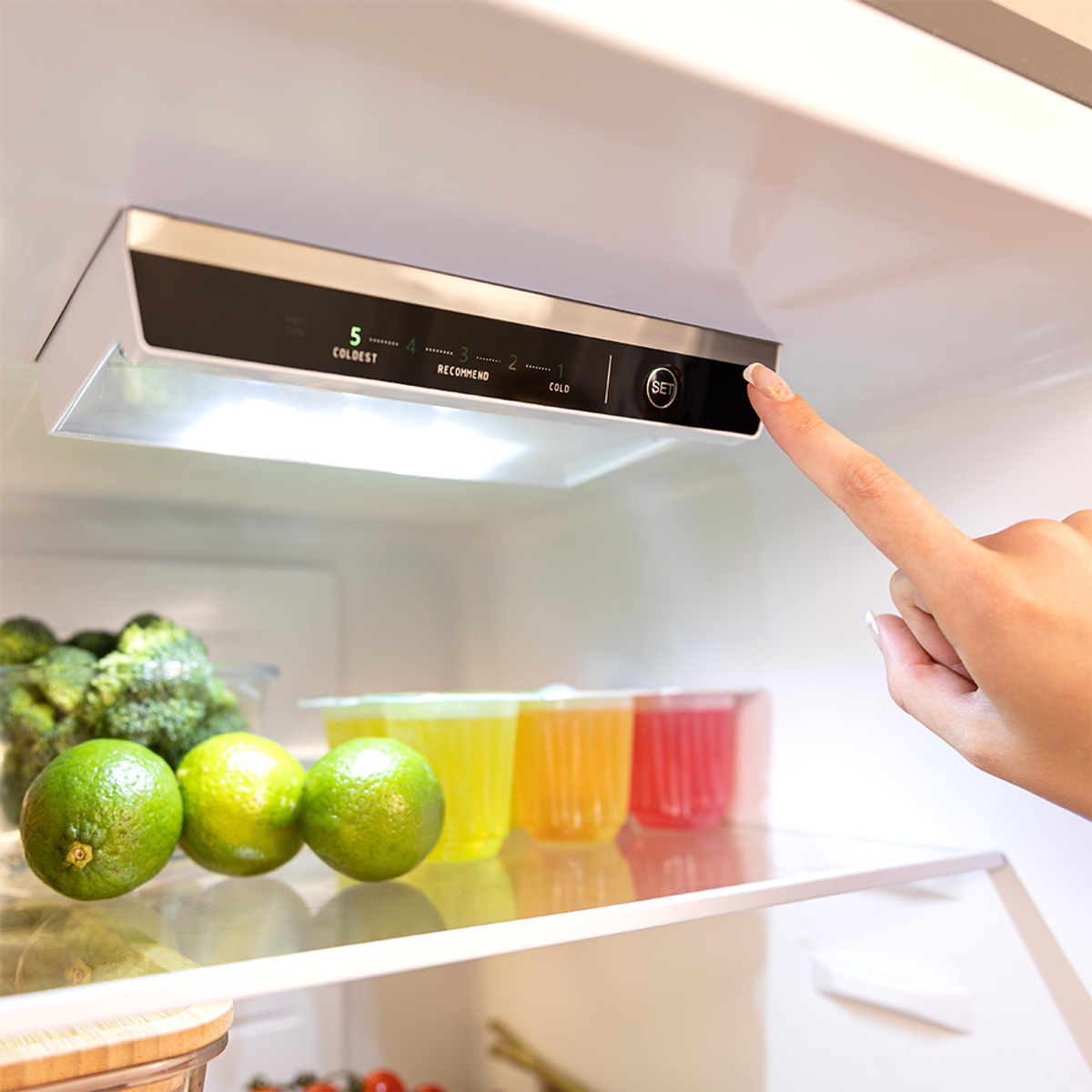 Mano che imposta la temperatura del frigorifero, lime e prodotti sugli scaffali visibili.