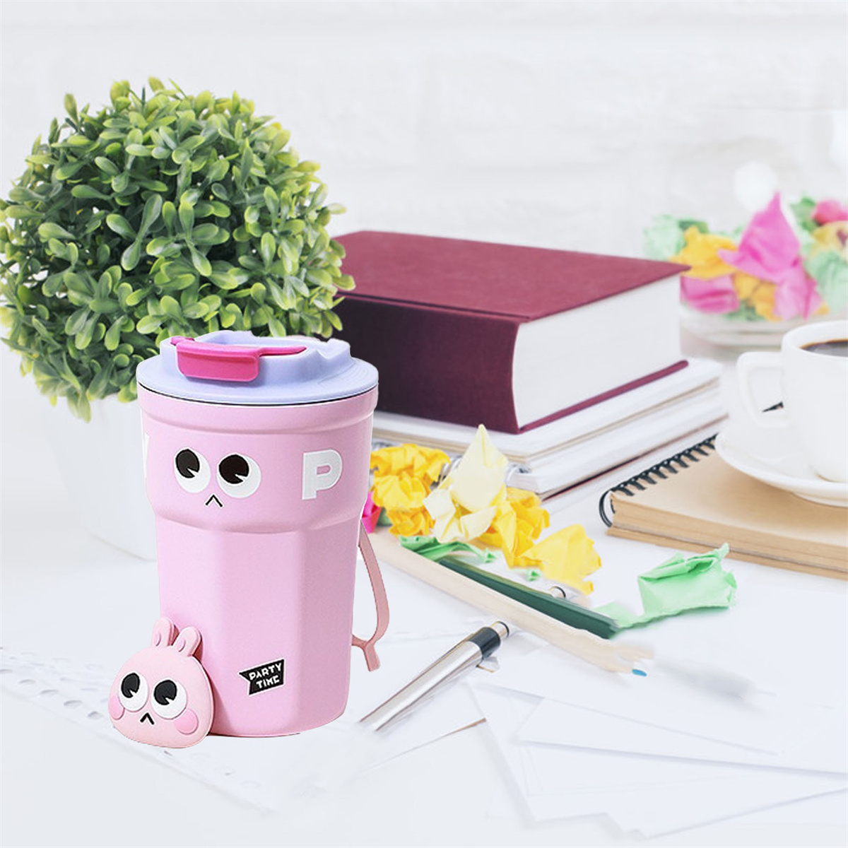 Ein rosa Becher mit einem Cartoon-Gesicht und einer Hasenfigur neben einer grünen Pflanze und Büchern auf einem Schreibtisch.