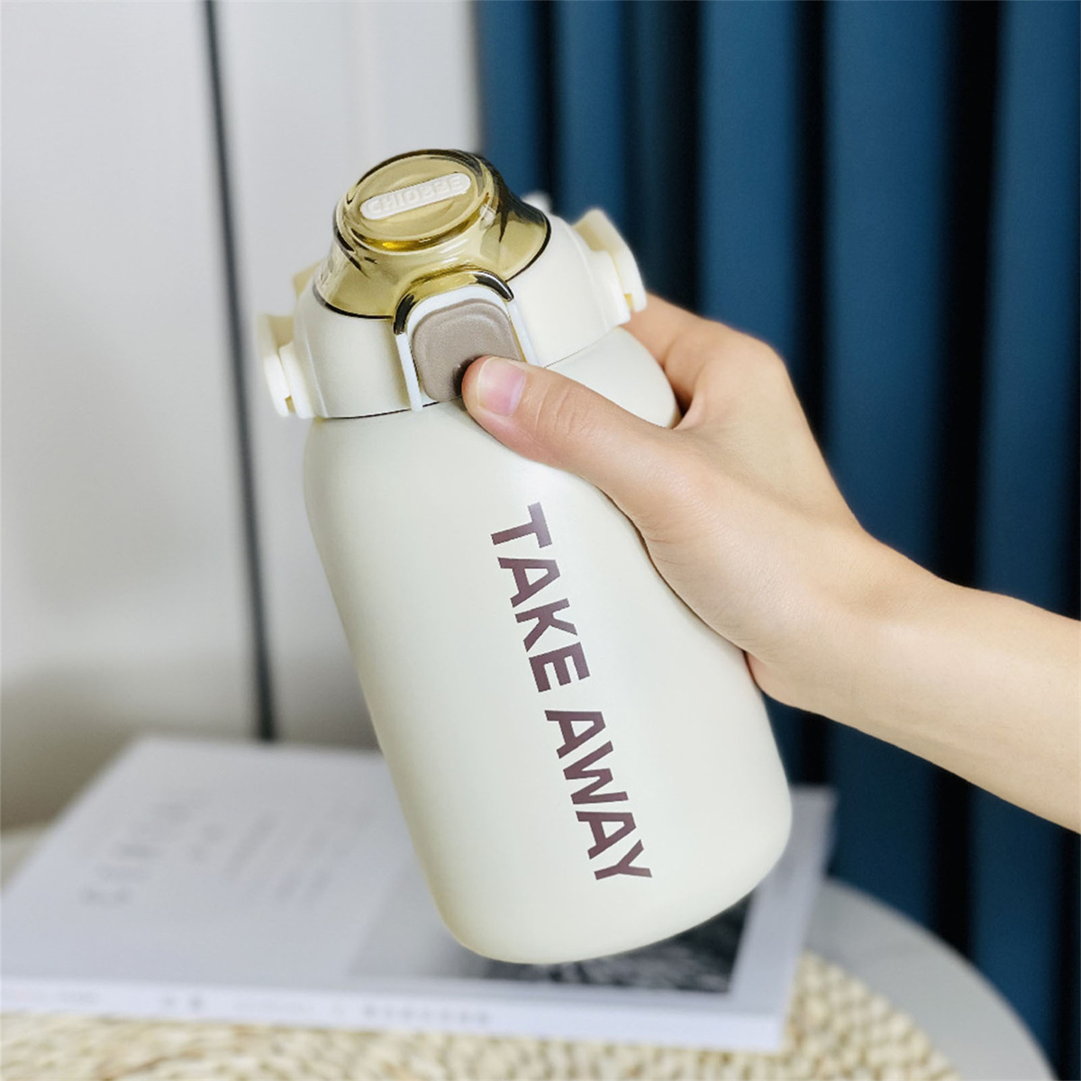 Cremefarbene Flasche mit Take Away-Etikett, gehalten von einer Hand. Goldfarbener Deckel.