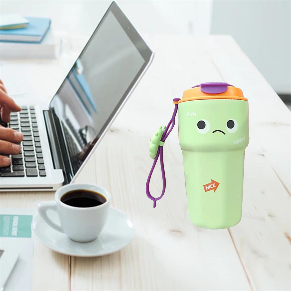 Ein grüner Becher mit Gesicht, eine Kaffeetasse und ein Laptop auf einem Schreibtisch.