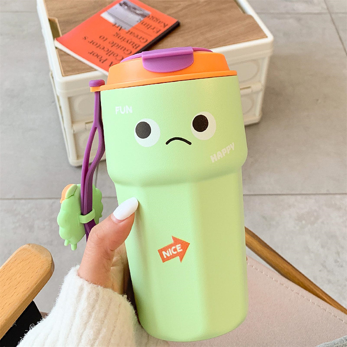 Eine grüne Wasserflasche mit Cartoon-Gesicht, lila Riemen und orangefarbenem Deckel. Von einer Hand gehalten.