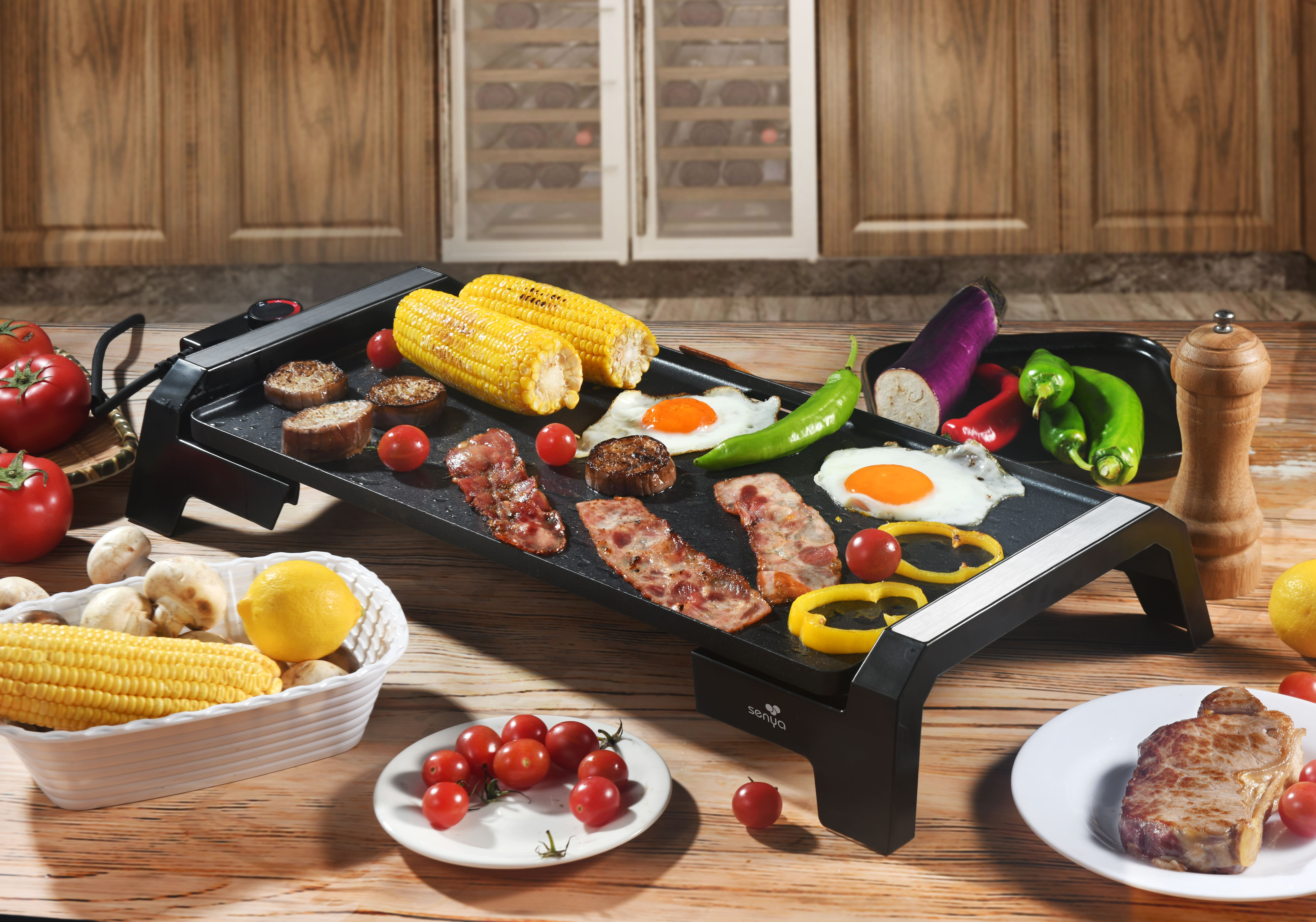Elektrogrill mit gekochten Speisen: Mais, Tomaten, Eier, Speck und Pilze. Holztisch.