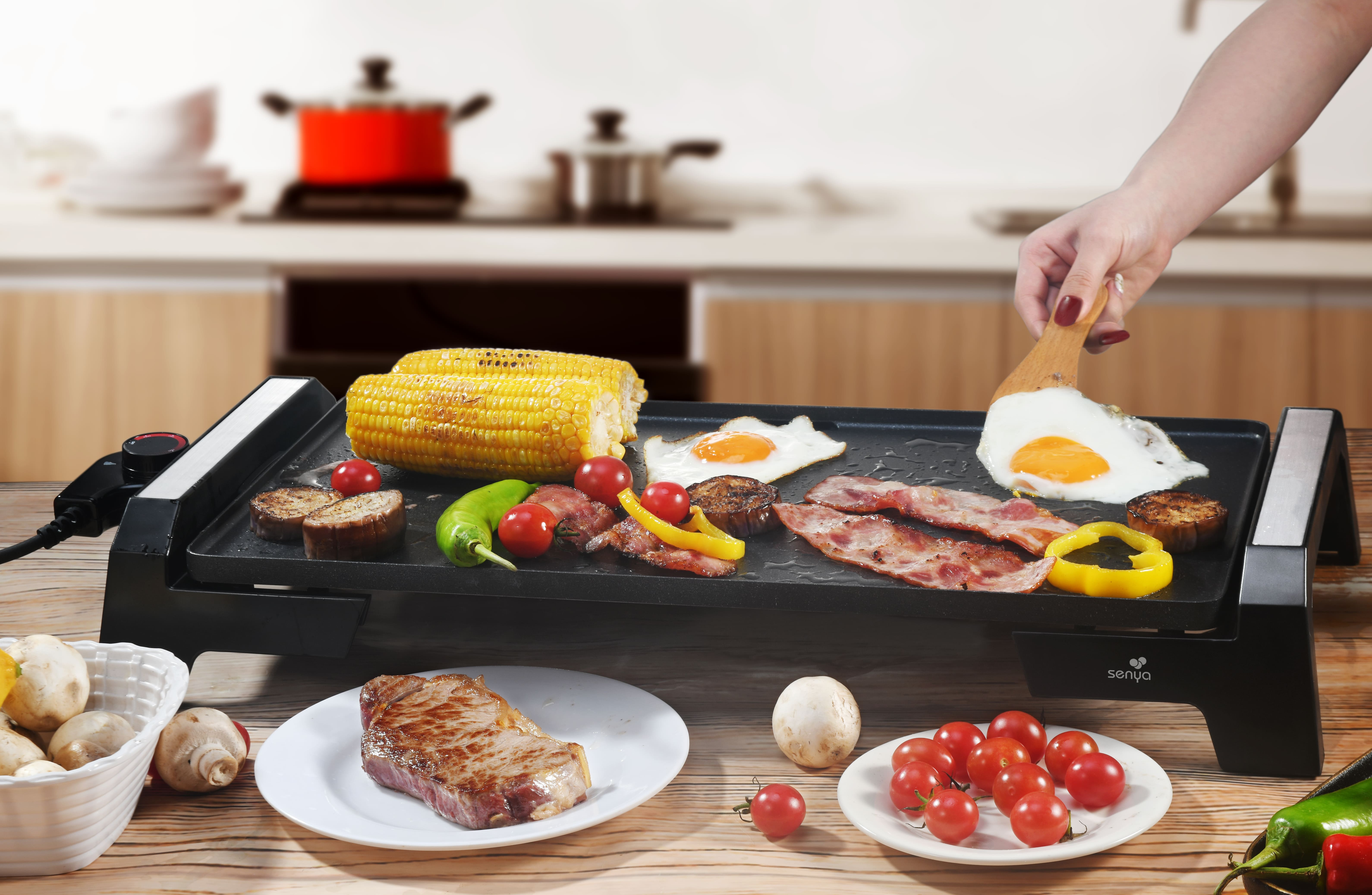 Eine Person kocht Essen auf einem elektrischen Grill. Enthält Mais, Eier, Speck und Tomaten.