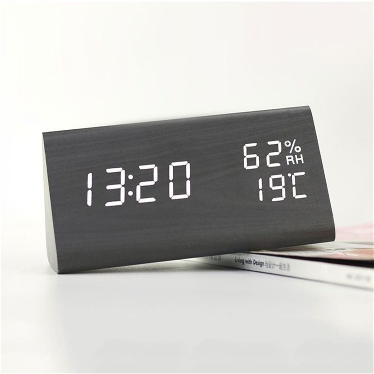 Eine schwarze Digitaluhr, die Zeit, Luftfeuchtigkeit und Temperatur auf einer weißen Oberfläche mit einem Magazin anzeigt.