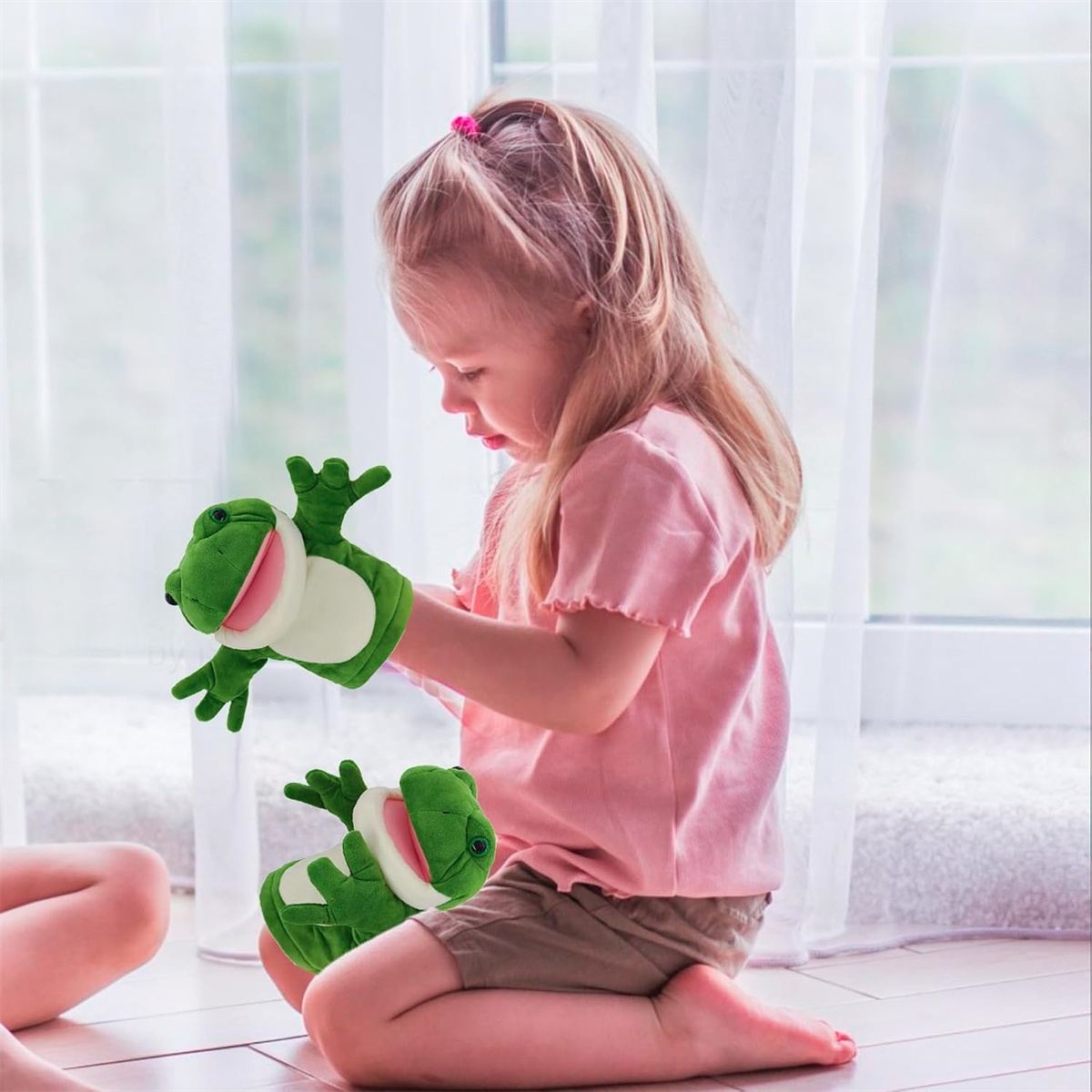 Ein junges Mädchen spielt mit Frosch-Handpuppen. Sie sitzt in der Nähe eines Fensters und trägt ein rosa Hemd.
