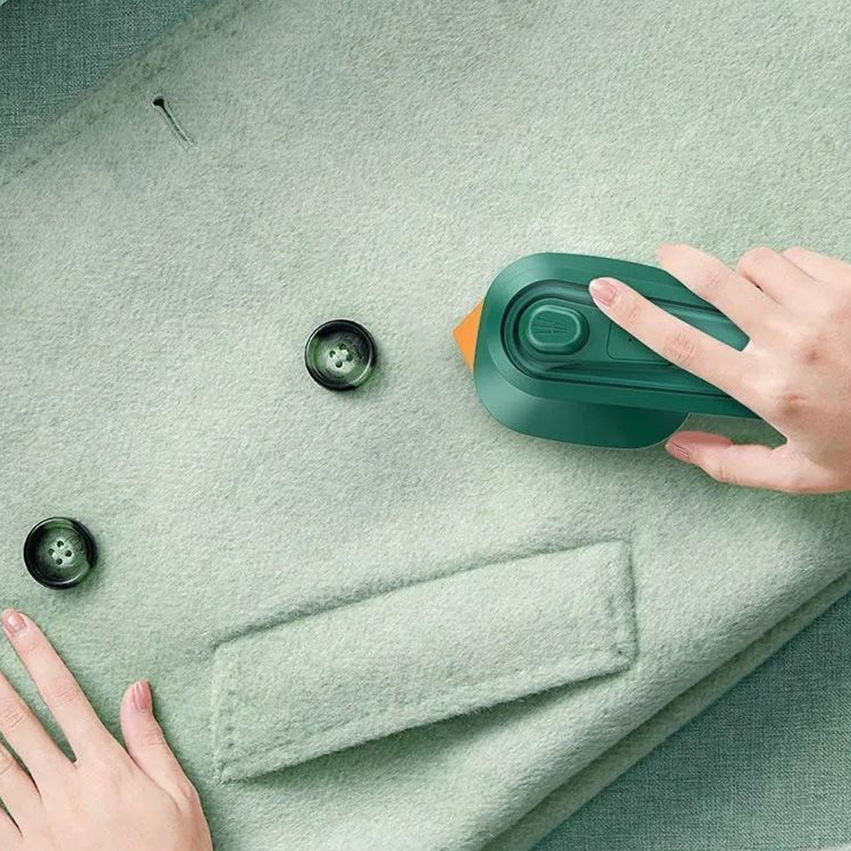 Person benutzt ein Mini-Bügeleisen auf einem grünen Mantel, in der Nähe von Knöpfen und Tasche.