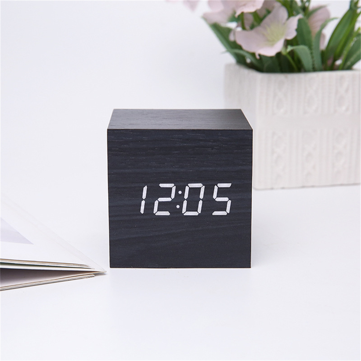 Eine schwarze Holzwürfeluhr, die 12:05 mit weißem Licht anzeigt, auf weißer Oberfläche.