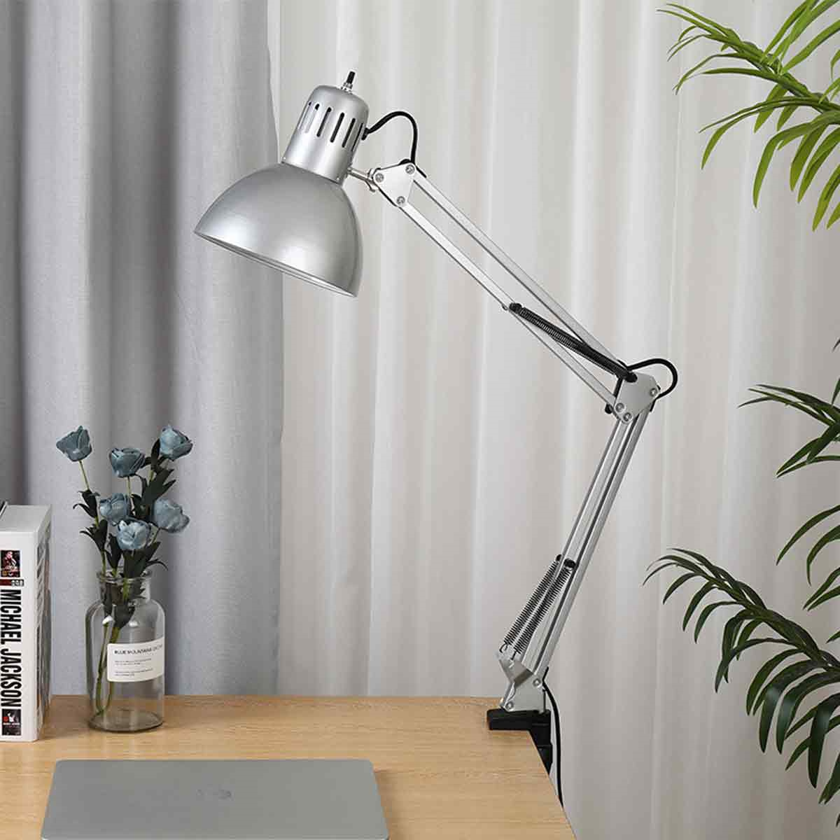 Eine silberne Schreibtischlampe steht auf einem Holztisch. Ein Laptop und Blumen sind sichtbar.