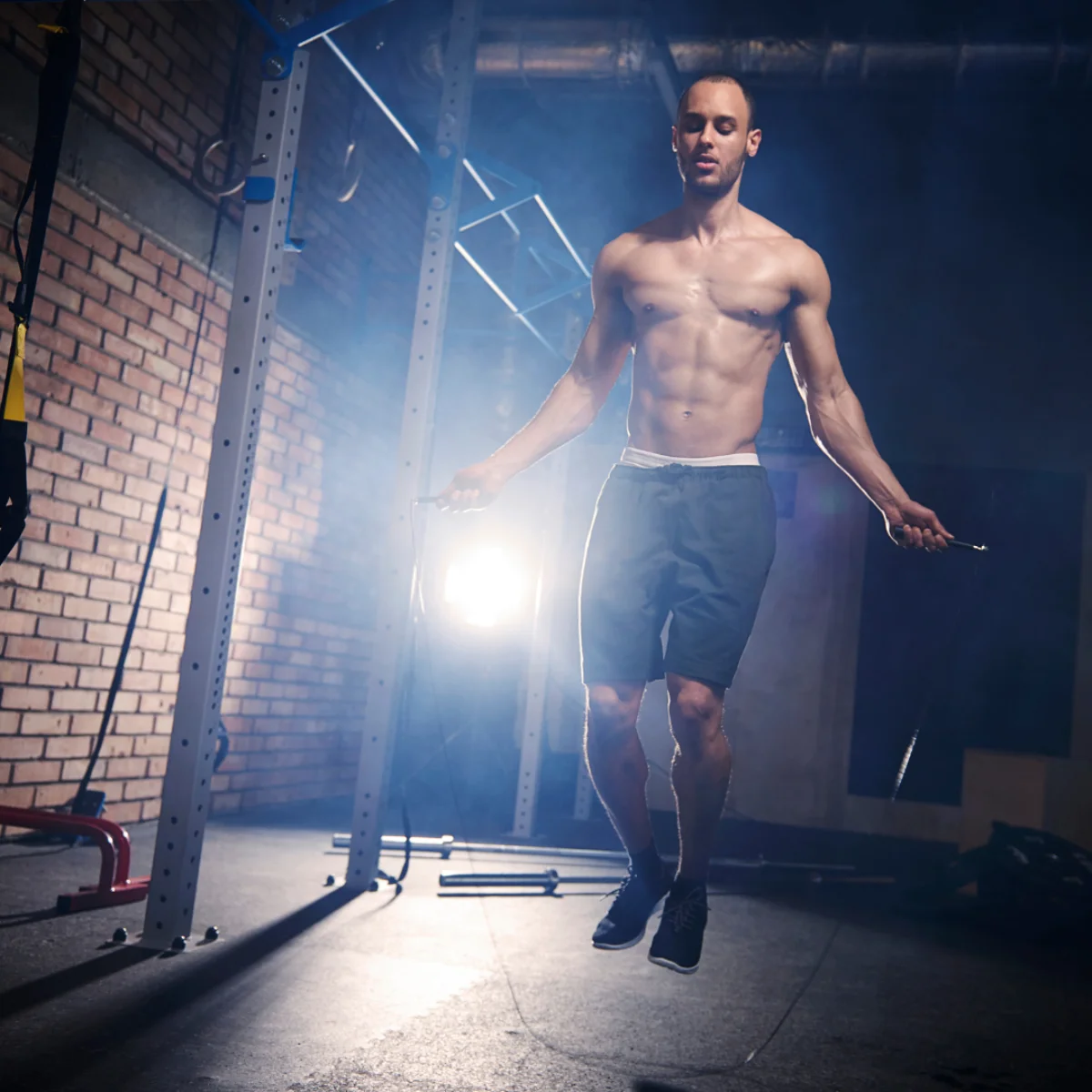 Ein Mann ohne Hemd springt in einem Fitnessstudio Seil, im Hintergrund Ziegelmauer und Geräte.