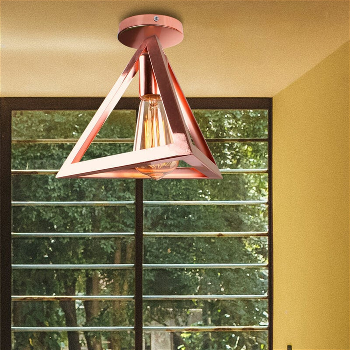 Roségoldene dreieckige Deckenleuchte mit Glühbirne, in der Nähe eines Fensters mit grünem Laub.